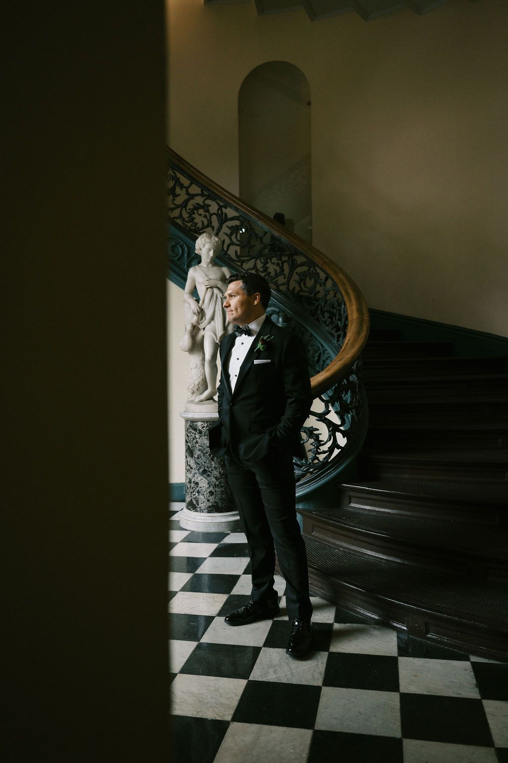 Dramatic portrait on checkered floor with ornate staircase in background.