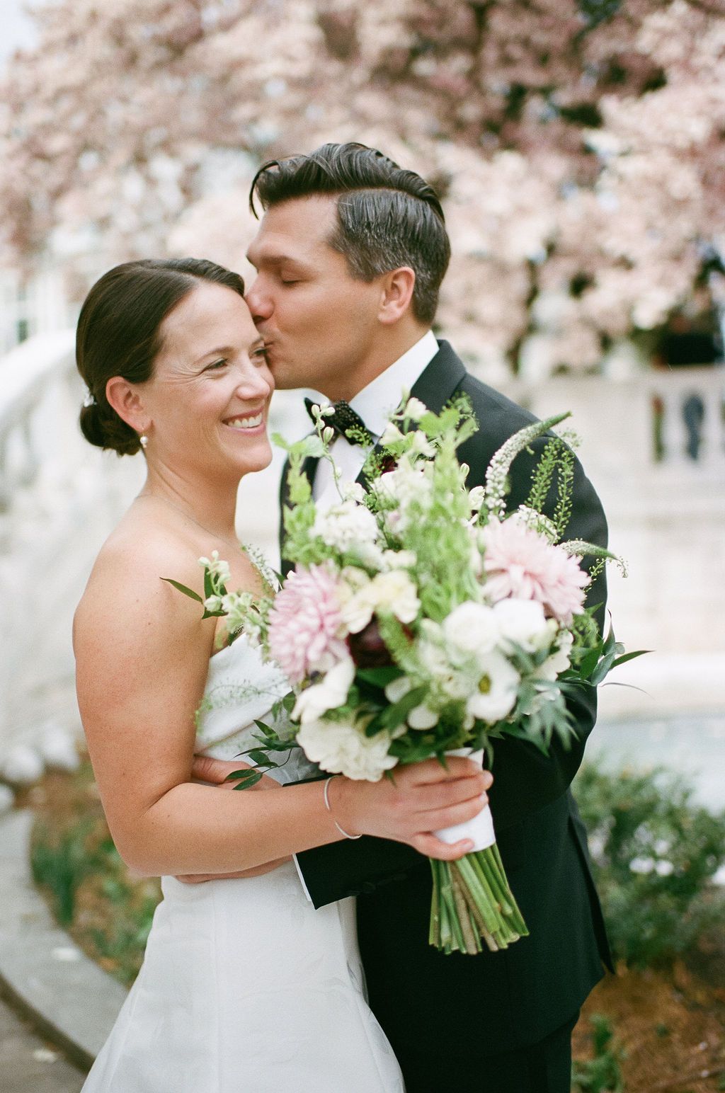 Joyful wedding moment captured under pink cherry blossoms with white bouquet.