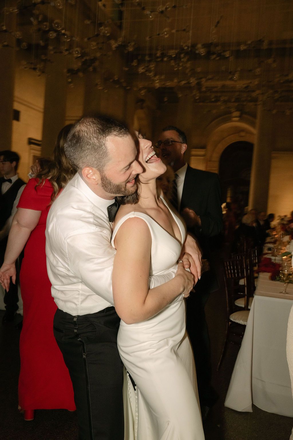 A couple shares a romantic dance and kiss at an elegant indoor reception venue with dim lighting.