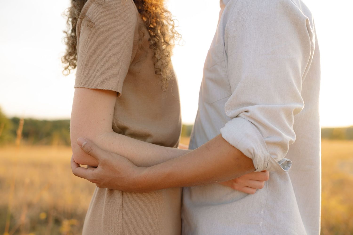 Close-up of couple embracing in sunlit field wearing light colored clothing.