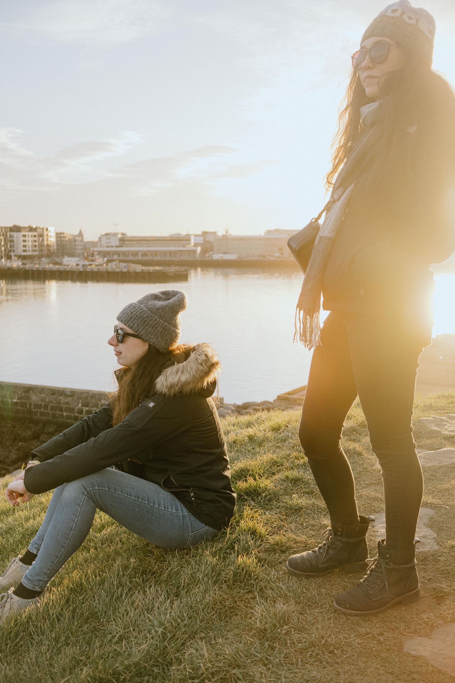 Two people enjoying sunset by waterfront on grassy slope.