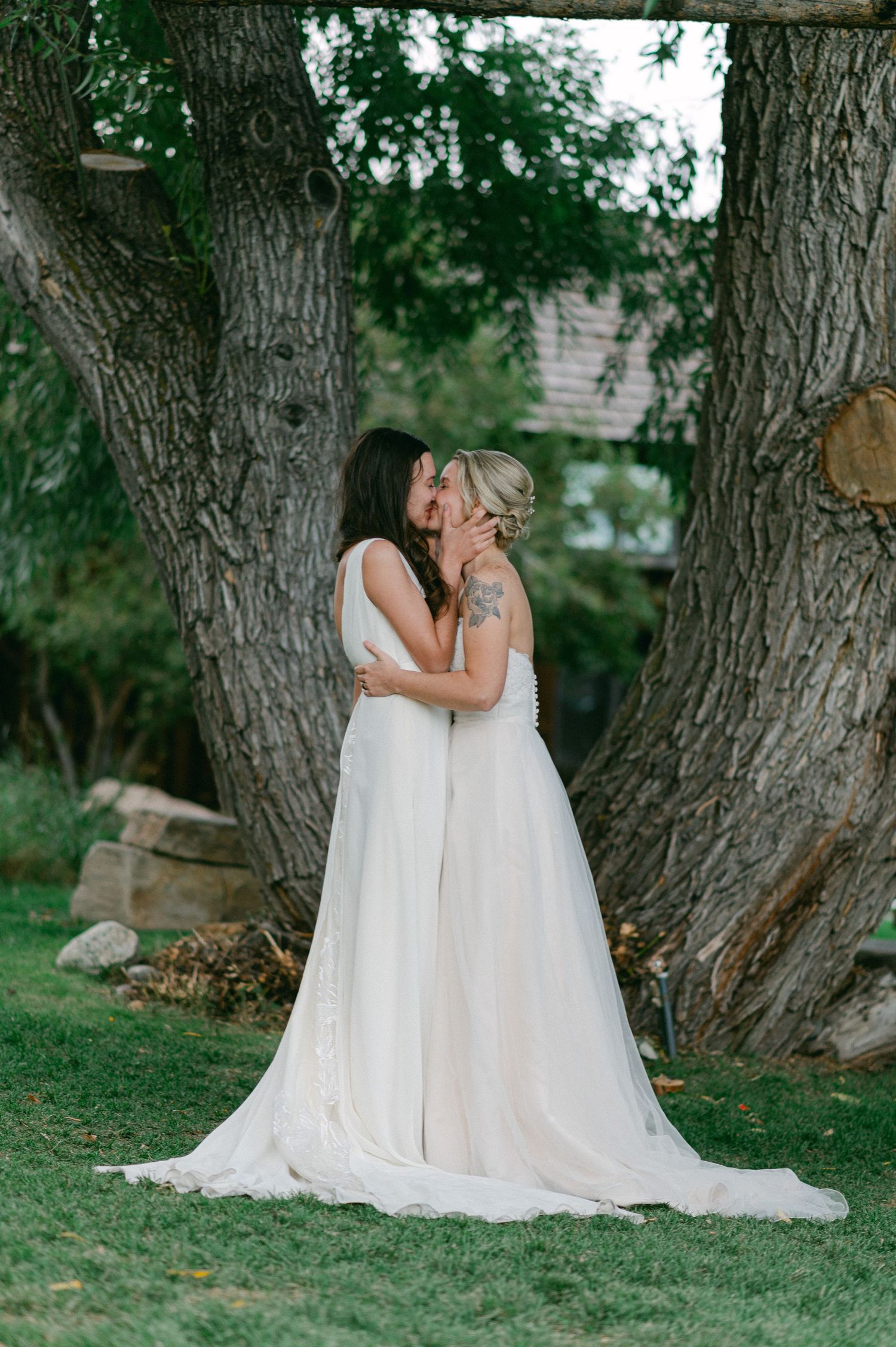 Two people in white wedding dresses share an intimate moment between large tree trunks in a garden setting.