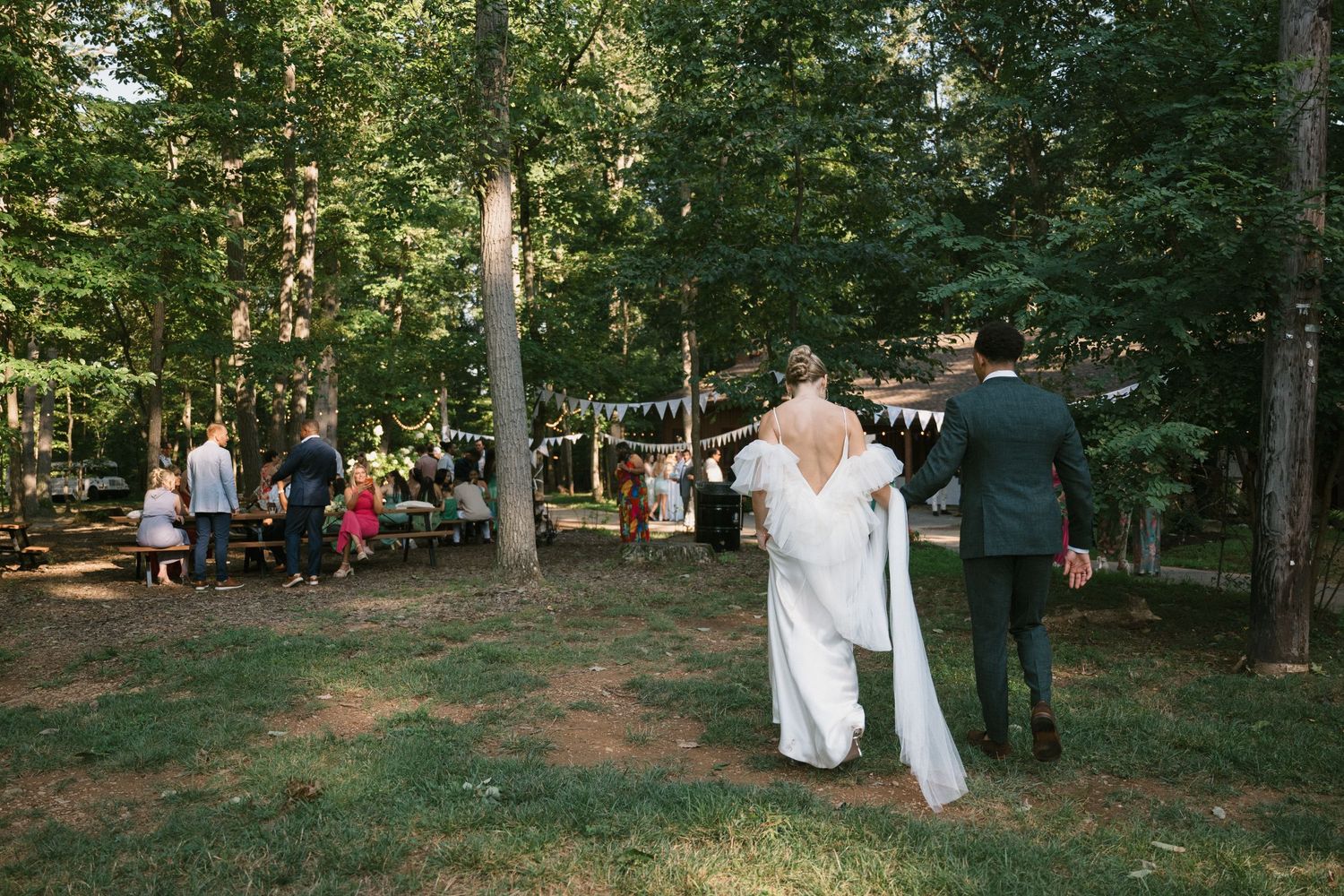 A couple walks away from their woodland wedding ceremony while guests mingle in the background.