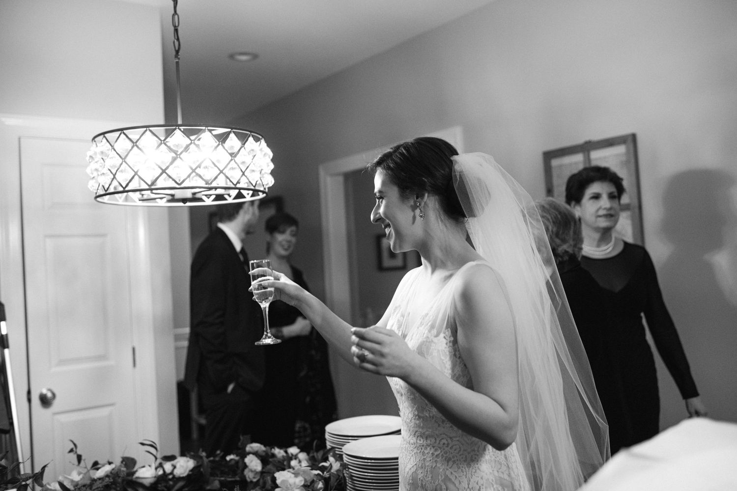 A bride in a white gown and veil stands indoors under an elegant chandelier lighting fixture during wedding preparations.
