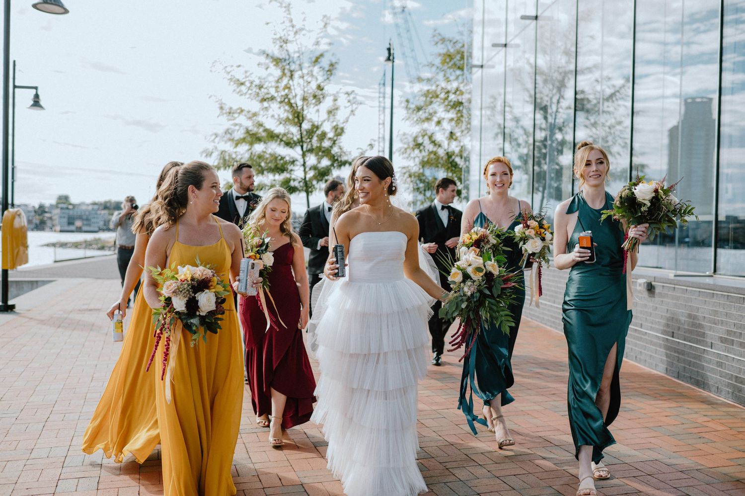 Wedding party walks along waterfront promenade with bridesmaids in colorful dresses.