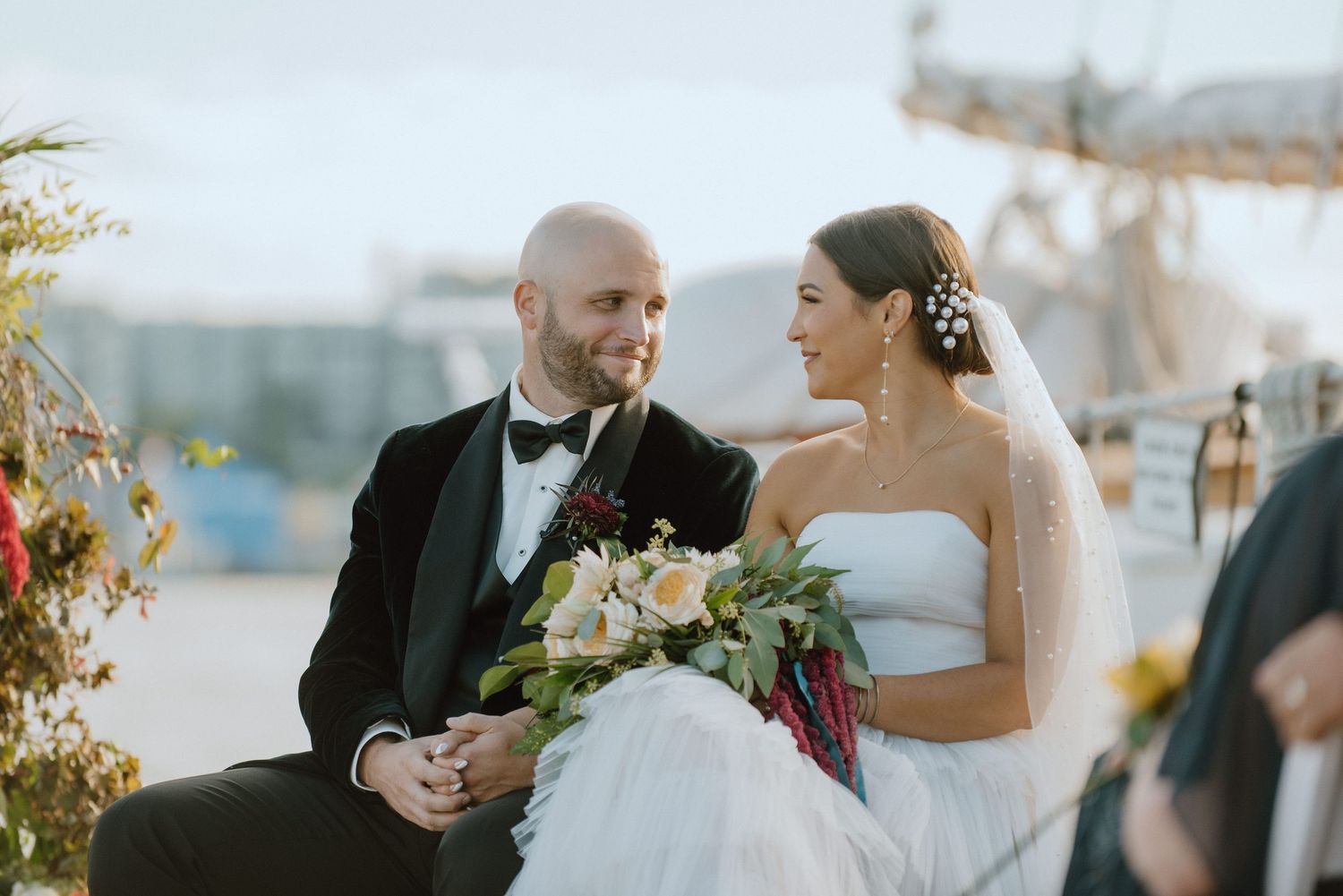 A couple sits together during their waterfront wedding ceremony with floral decorations visible.