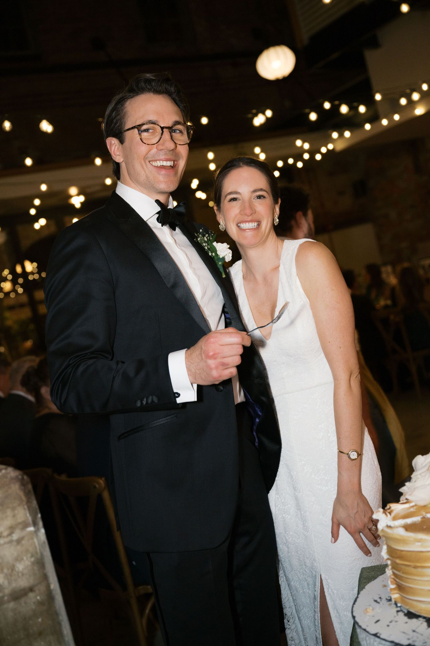 A couple at their wedding reception laughs together beside their cake under twinkling string lights.