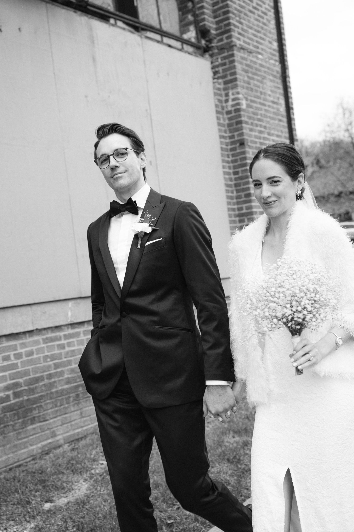 A black and white photo of a bride and groom standing together by a brick wall, with the bride holding a small bouquet.