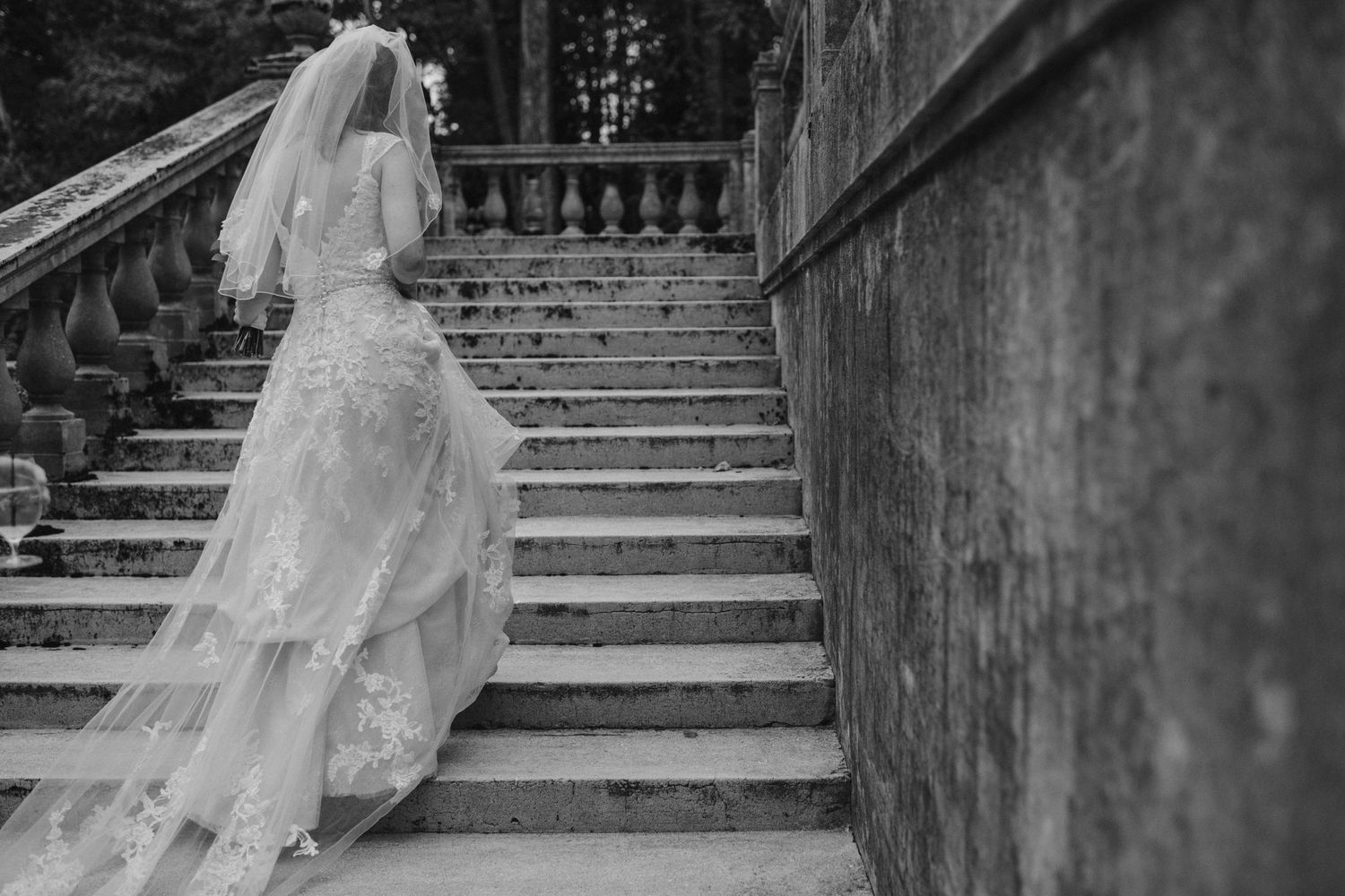 Bride in flowing lace gown ascends stone staircase in dramatic black and white photography.