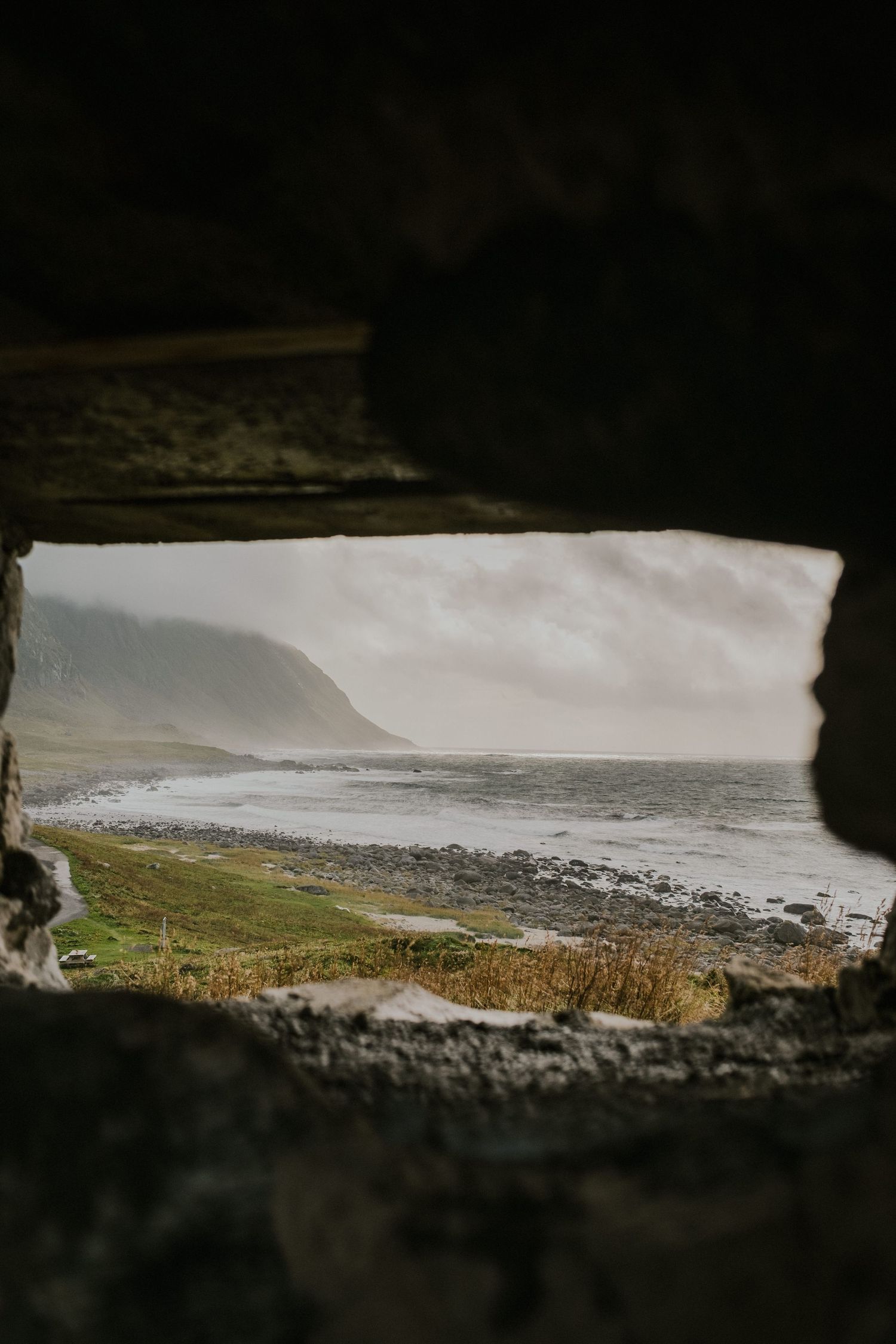 Dramatic coastal view framed through stone window opening with rocky beach and misty cliffs.
