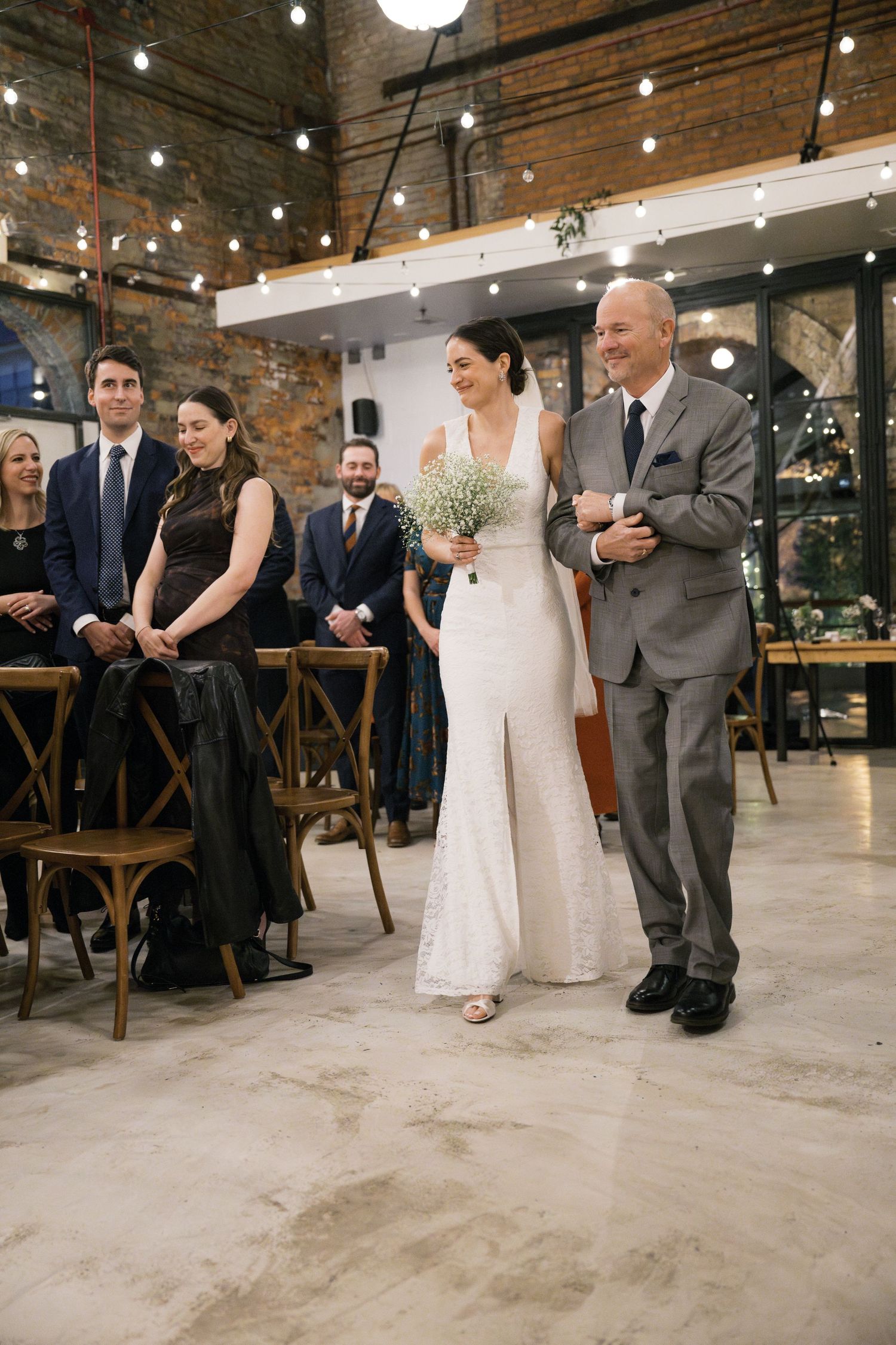 Wedding ceremony in a rustic industrial venue with exposed brick walls and string lights overhead during a processional moment.