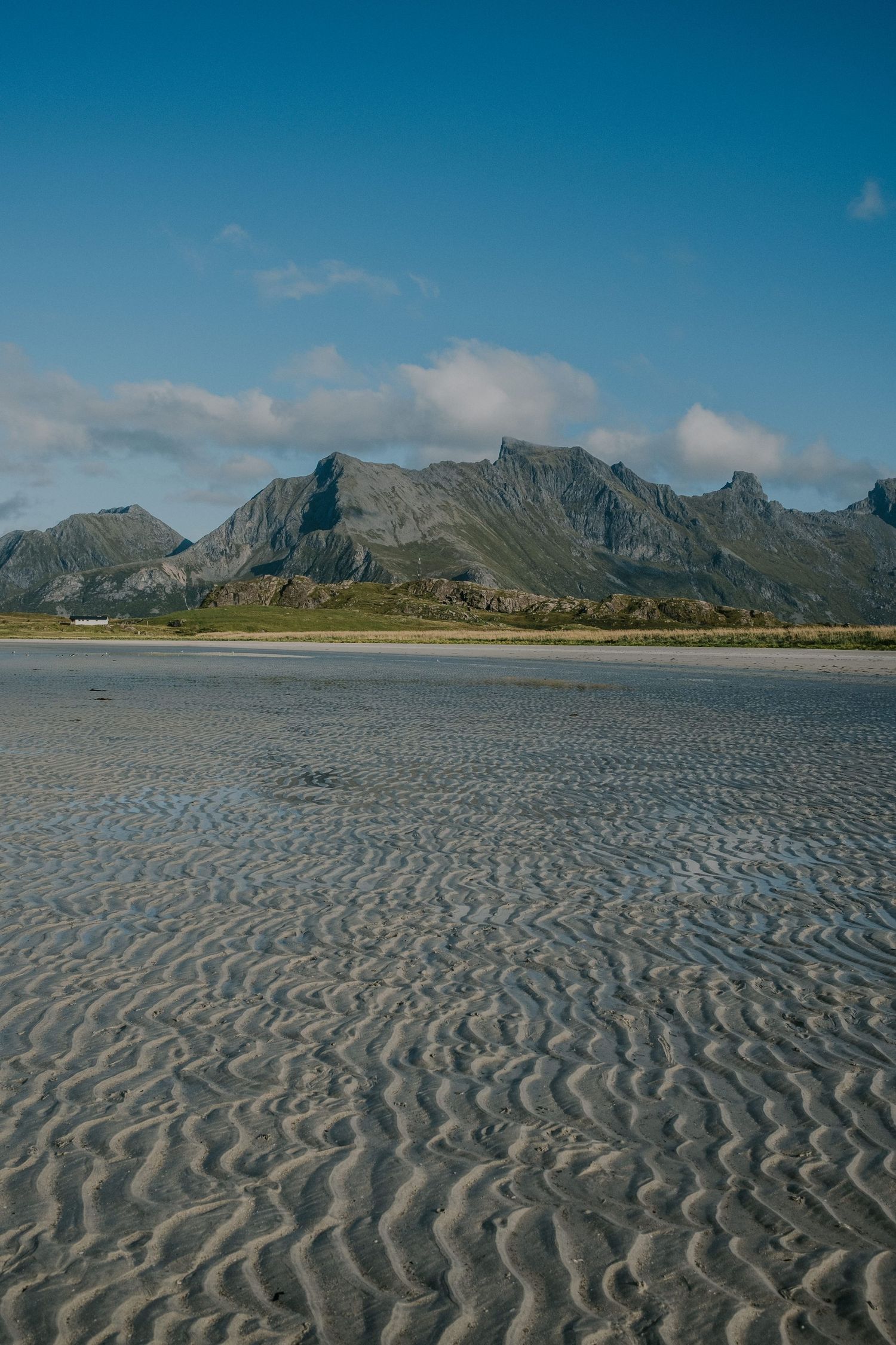 Rippled sand patterns on beach with majestic mountain range in background under blue sky.