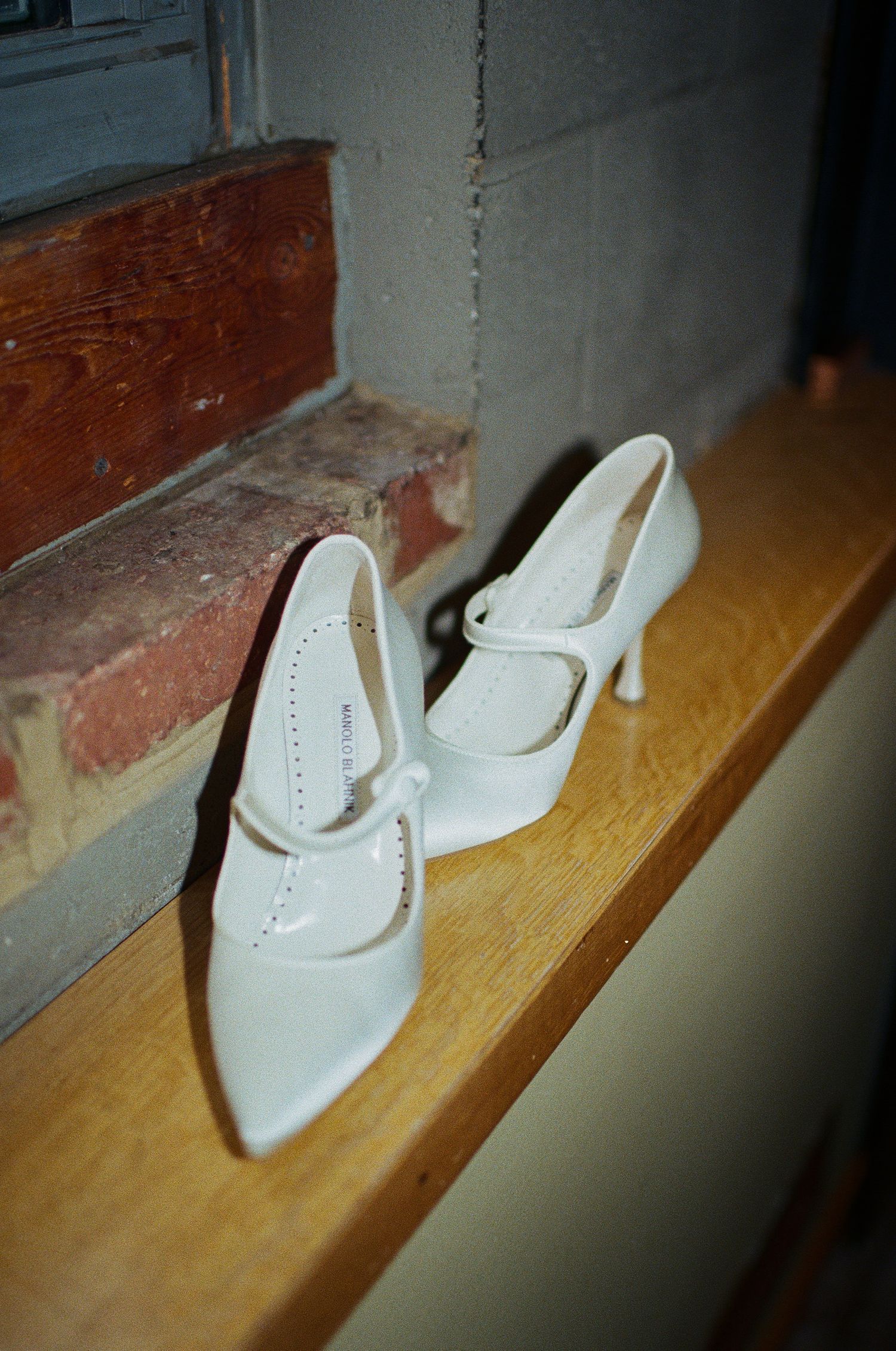 Elegant white vintage-style wedding shoes displayed on a wooden windowsill against brick and painted walls.