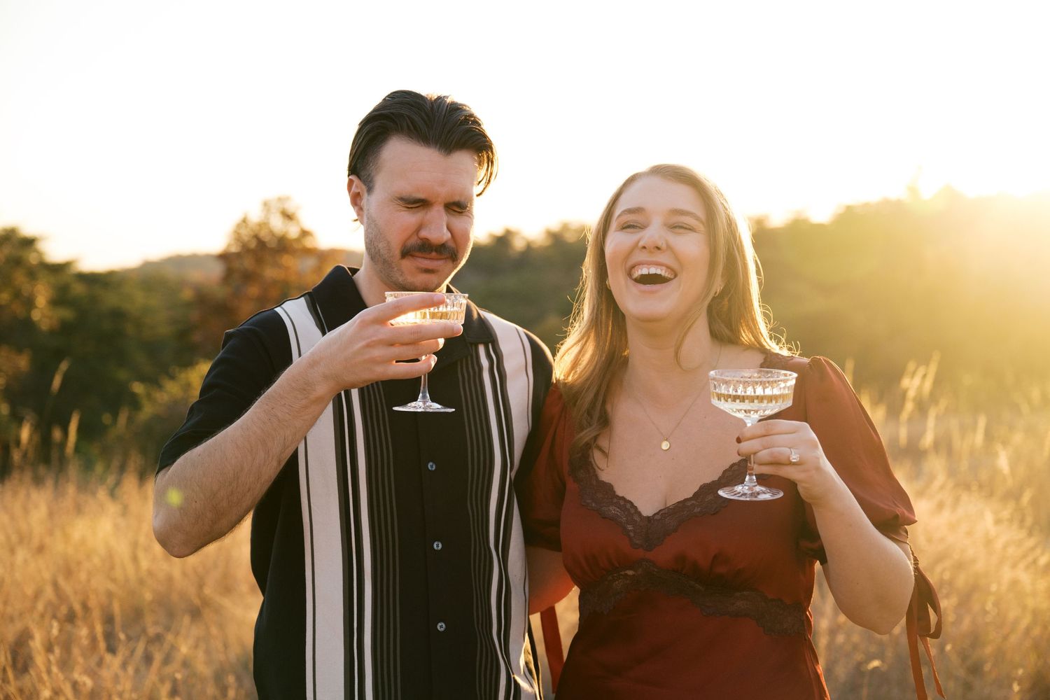A couple shares wine and laughter in a sun-drenched field during golden hour.
