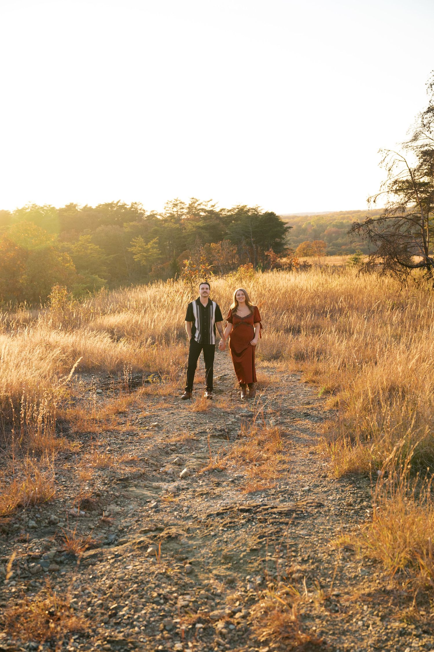 A couple stands together on a rocky path through golden grass during sunset with evergreen trees in background.