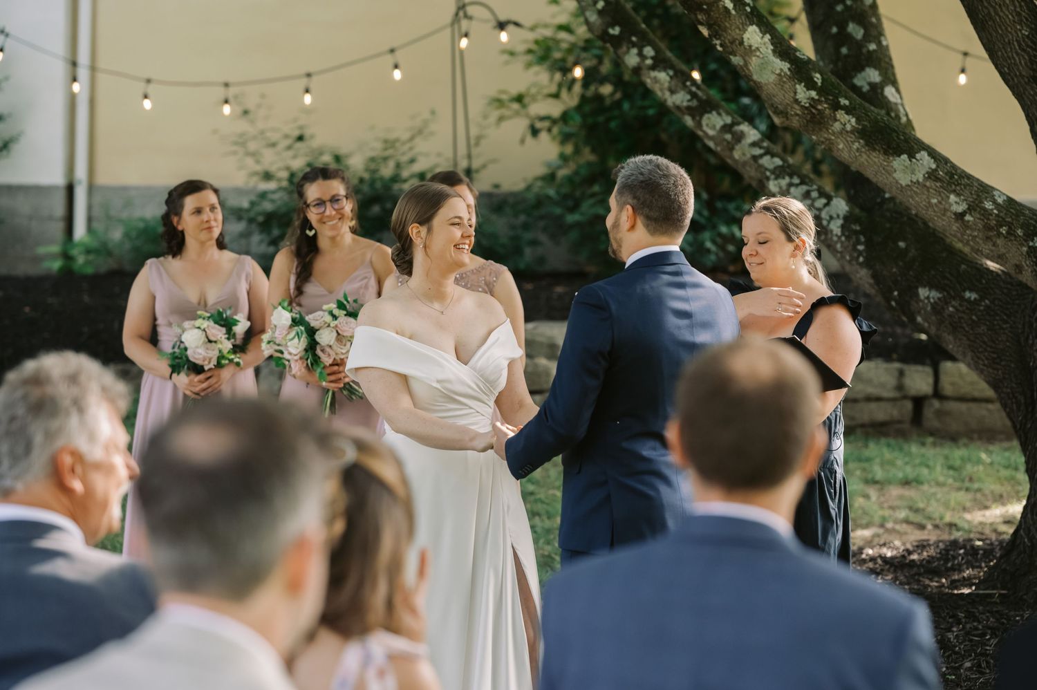 Wedding ceremony taking place under large trees with wooden chairs and pink parasol decorations.