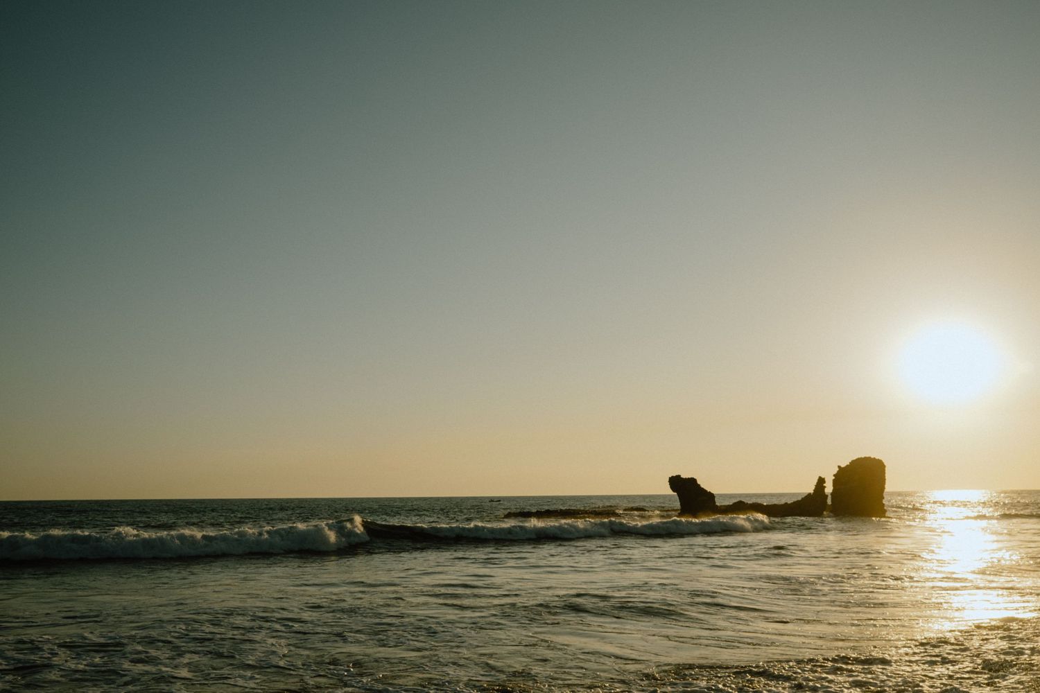 Sunset over the ocean with rocky formations silhouetted against the orange sky.