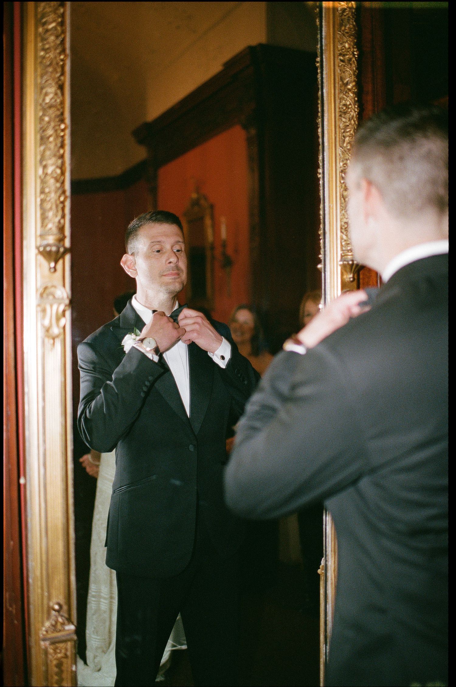A man adjusts his bowtie while looking in an ornate gold-framed mirror.