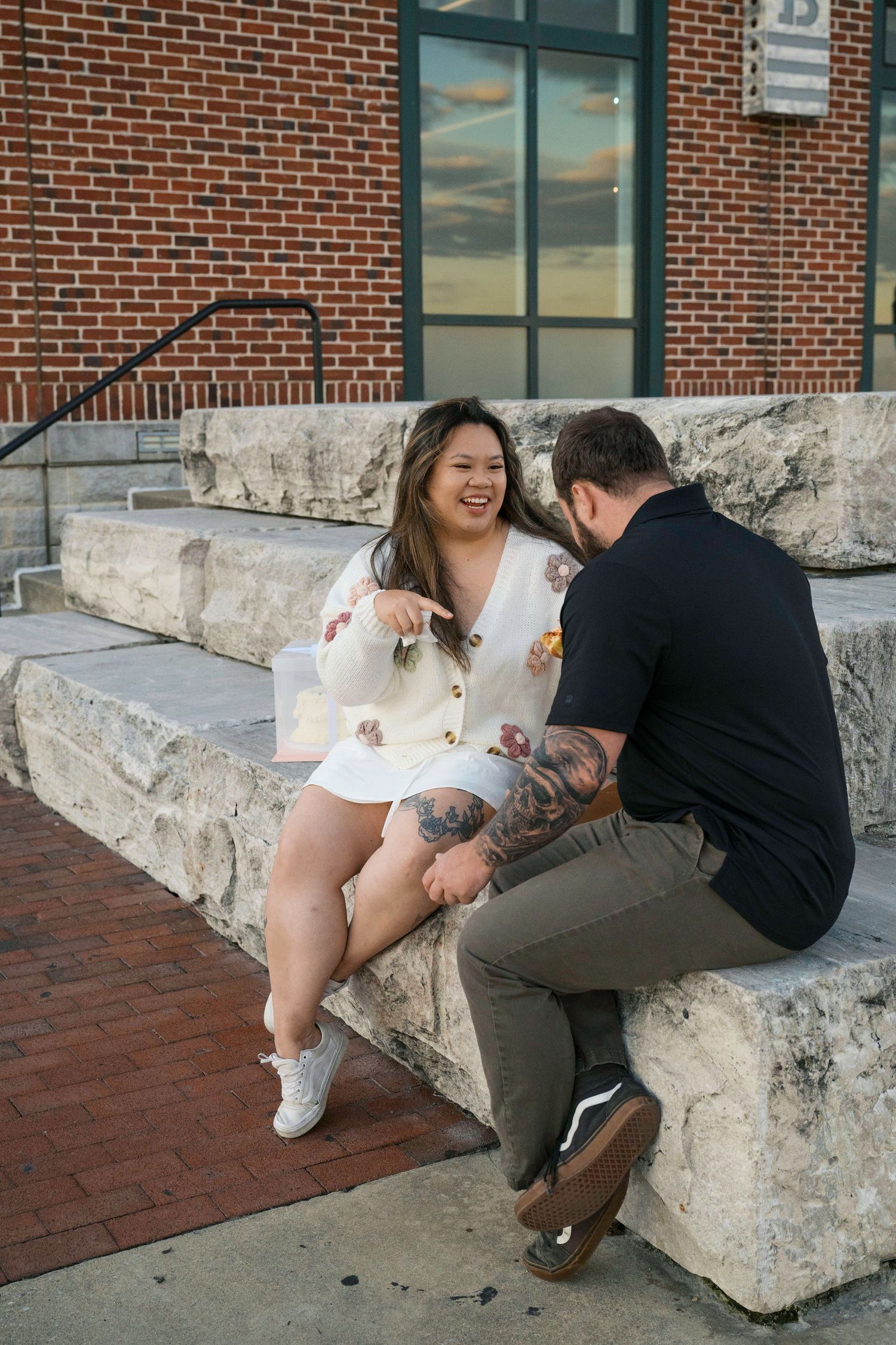 A couple shares a tender moment sitting together on concrete steps in front of a brick building at sunset.