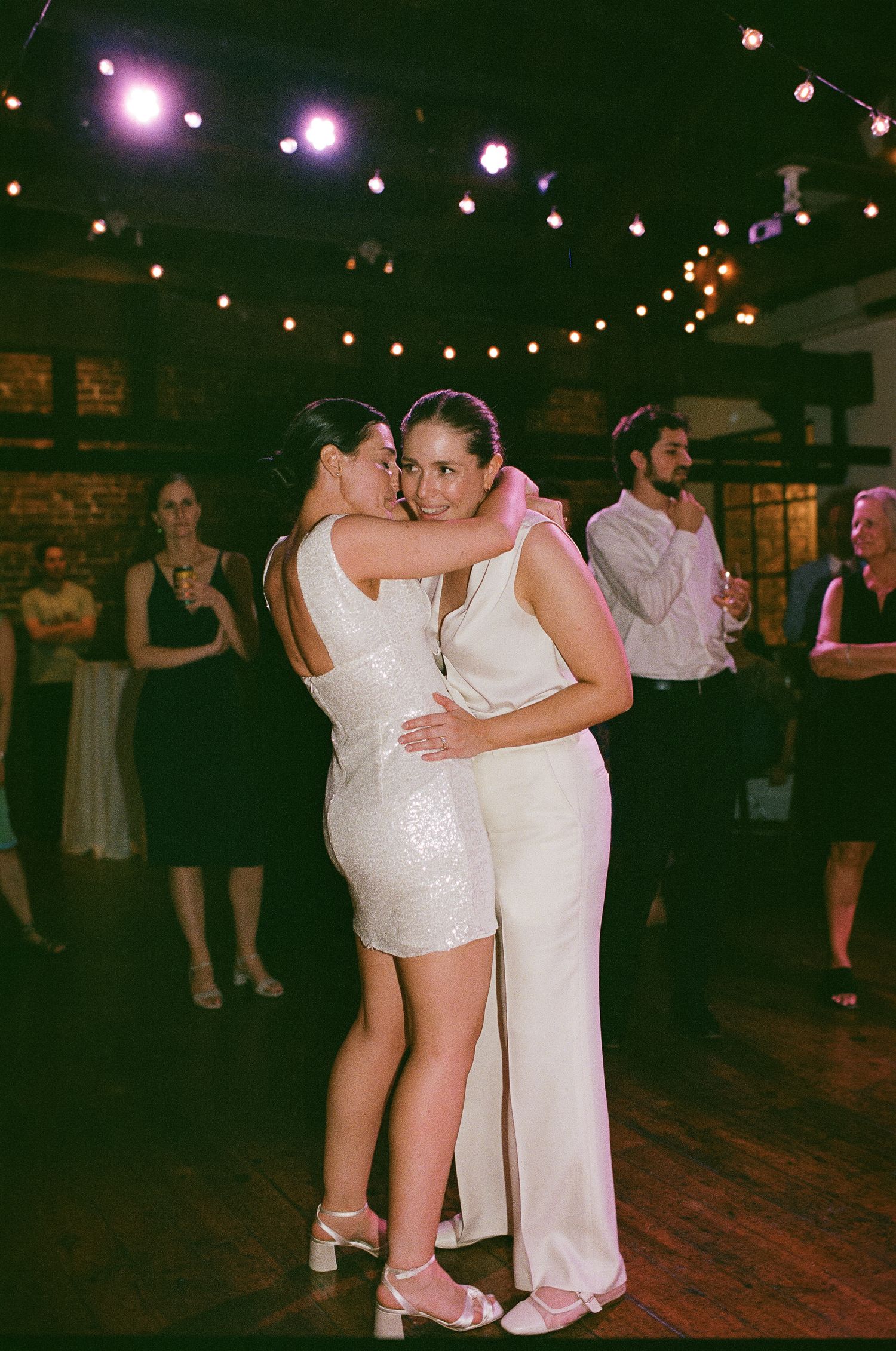 Dancing couple embrace at a wedding reception under twinkling string lights on a wooden dance floor.
