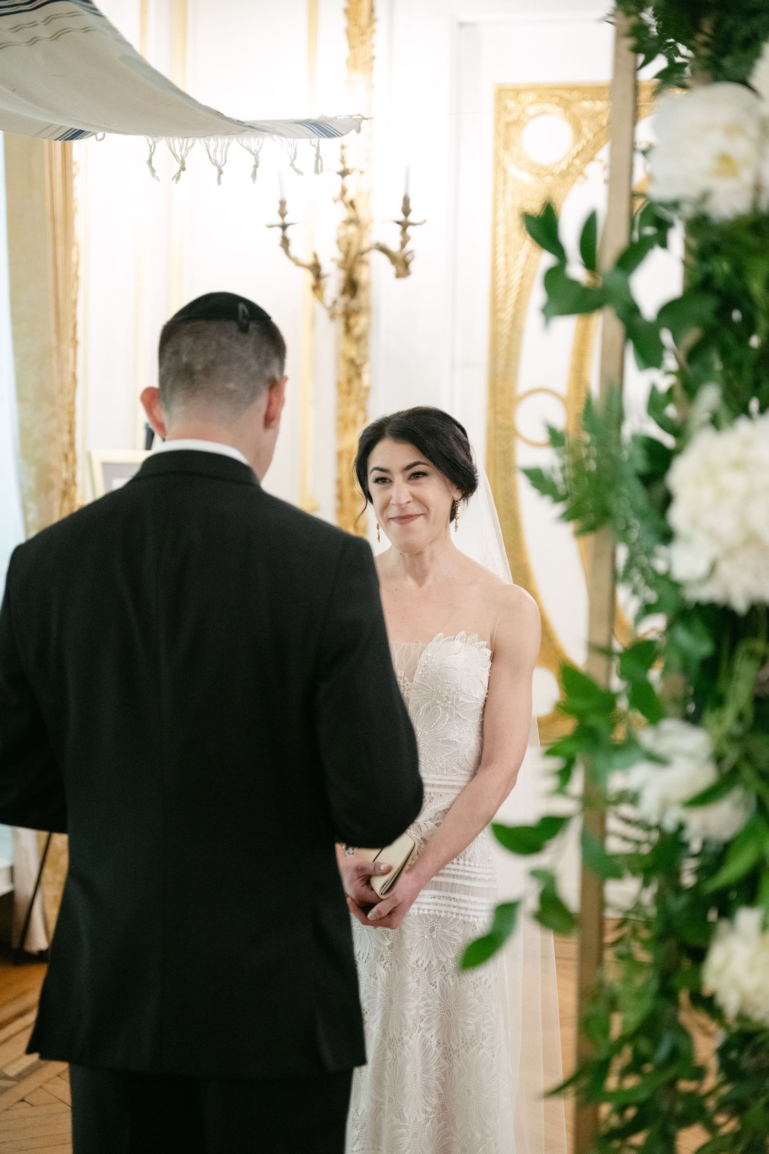 A tender wedding ceremony moment captured in an elegant room with ornate decor and white floral arrangements.