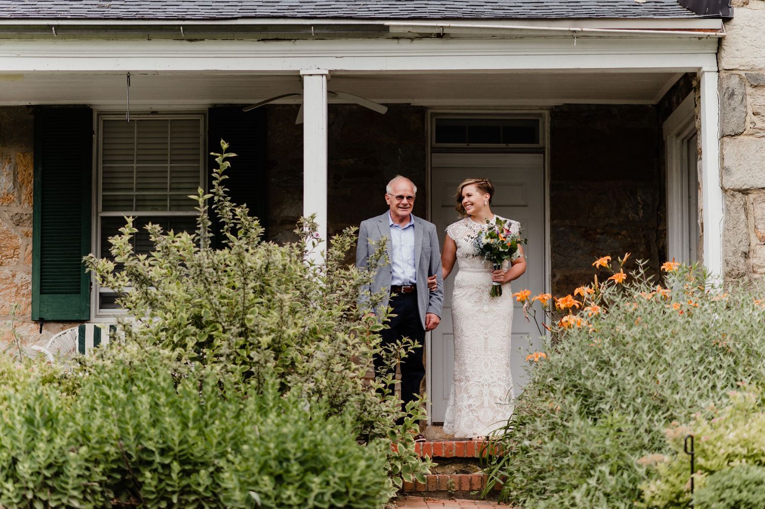 Couple standing on front porch of stone house with blooming garden.