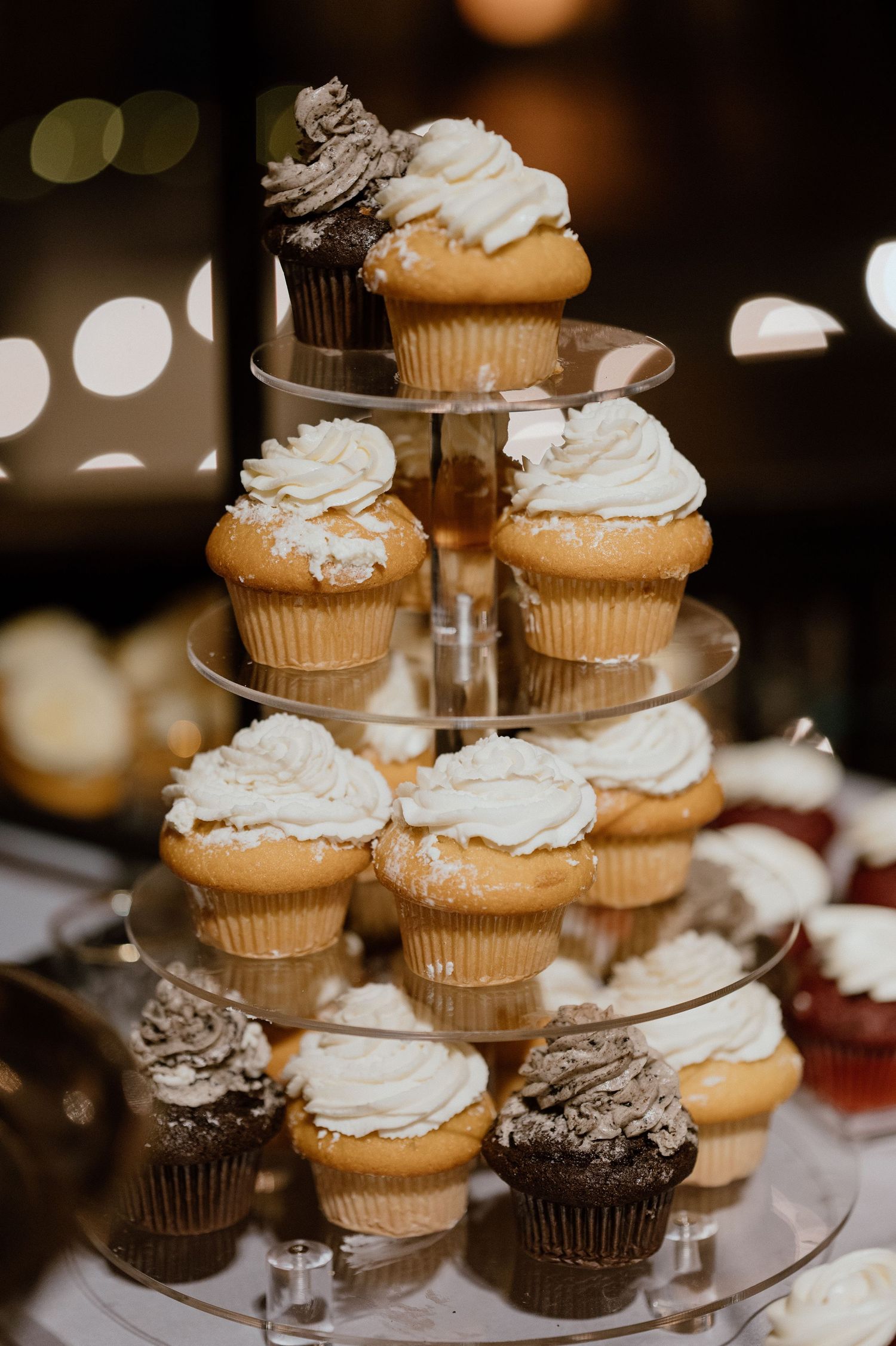 Tiered display of vanilla and chocolate cupcakes with swirled white frosting at a wedding dessert table.