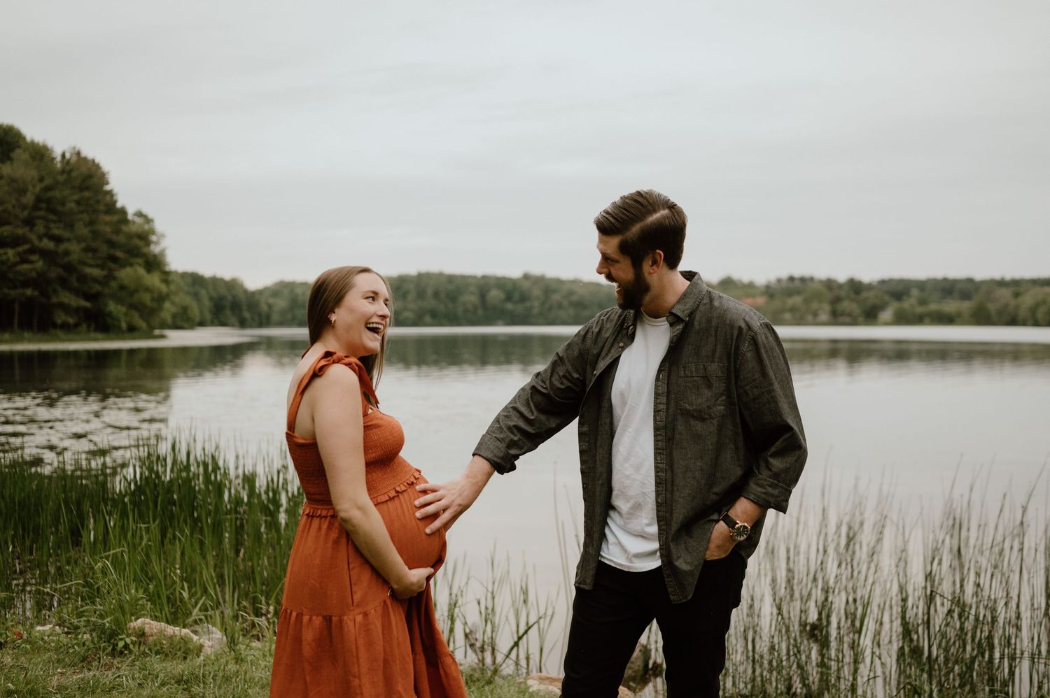 A couple shares a tender moment by a lake during their maternity photoshoot.