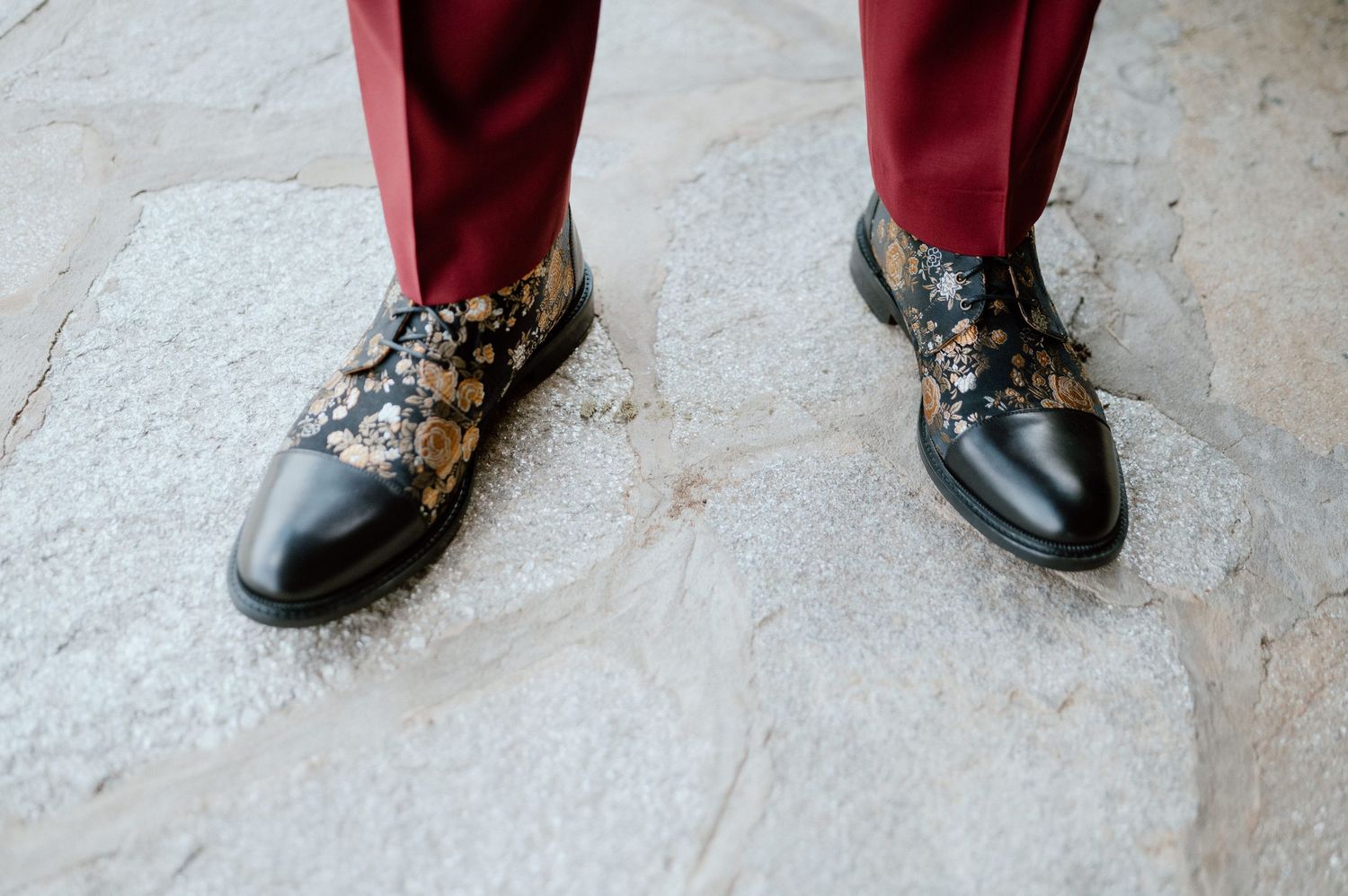 Dress shoes with decorative floral pattern worn with burgundy pants.