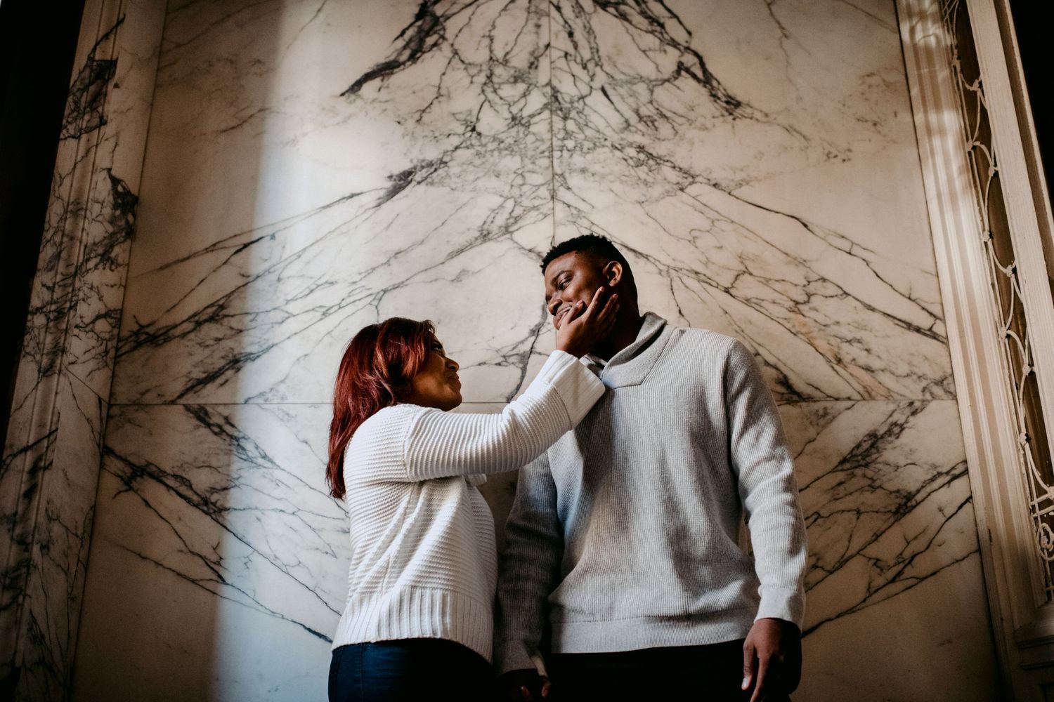 A romantic couple in white sweaters share an intimate moment against a marble wall backdrop.