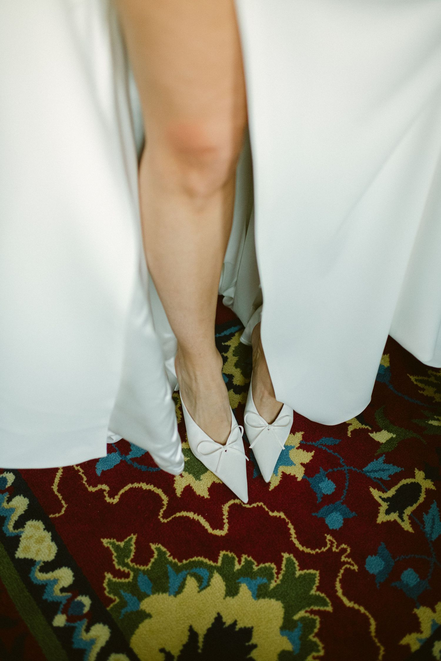 Close-up of white bridal shoes on an ornate floral carpet during wedding preparations.
