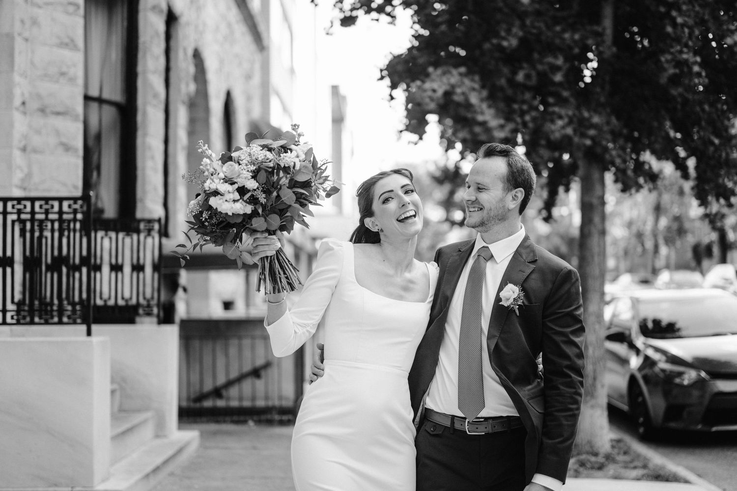 Black and white candid wedding photo showing couple laughing together on city street.