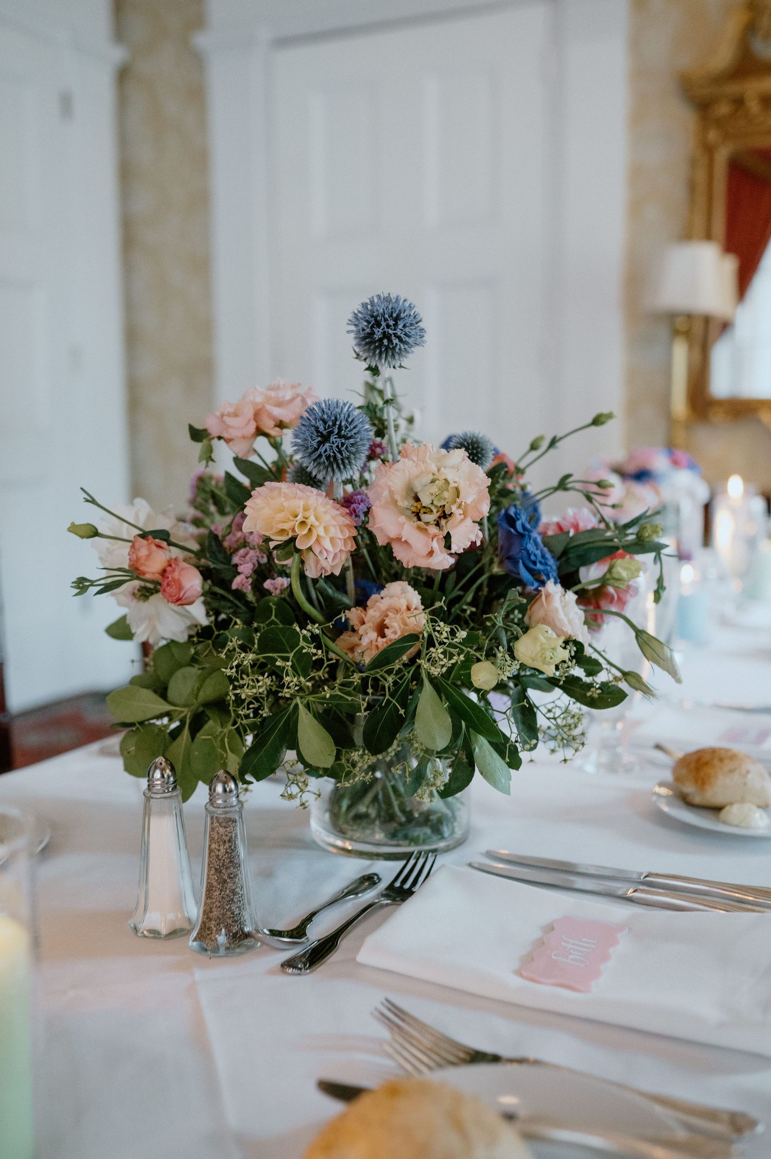 Romantic floral centerpiece with blue thistles, pink roses and greenery on white tablecloth.