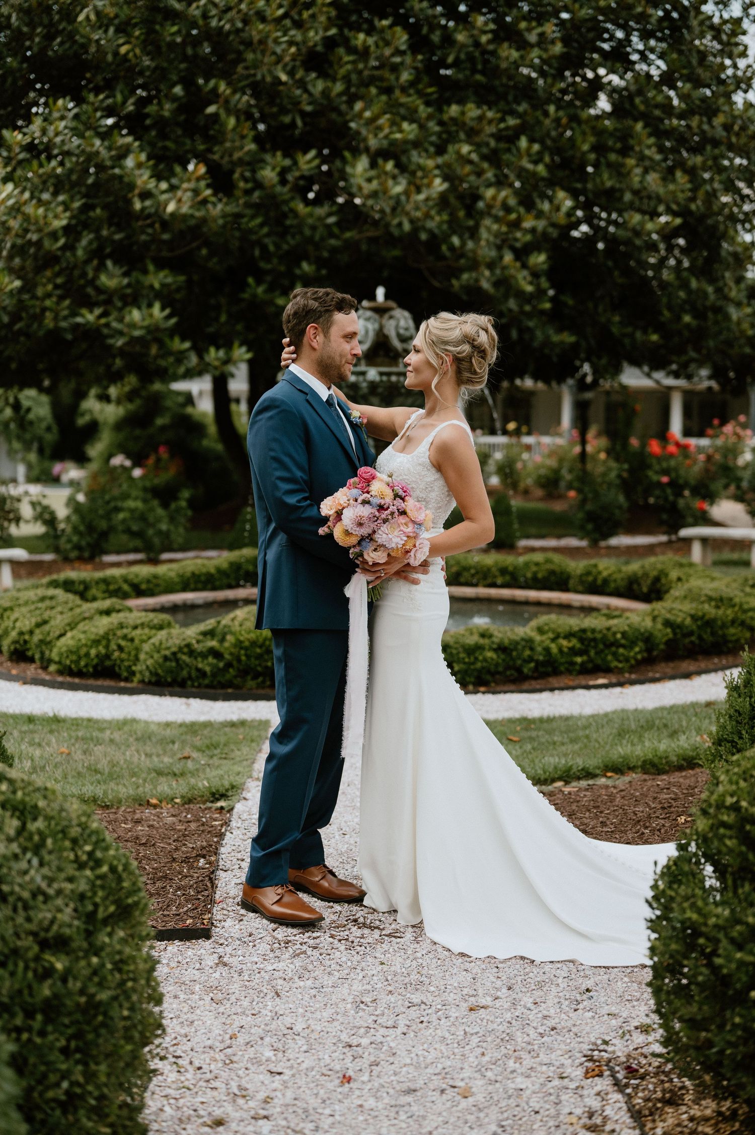 Elegant wedding couple poses in formal garden with pink bouquet and flowing white dress.