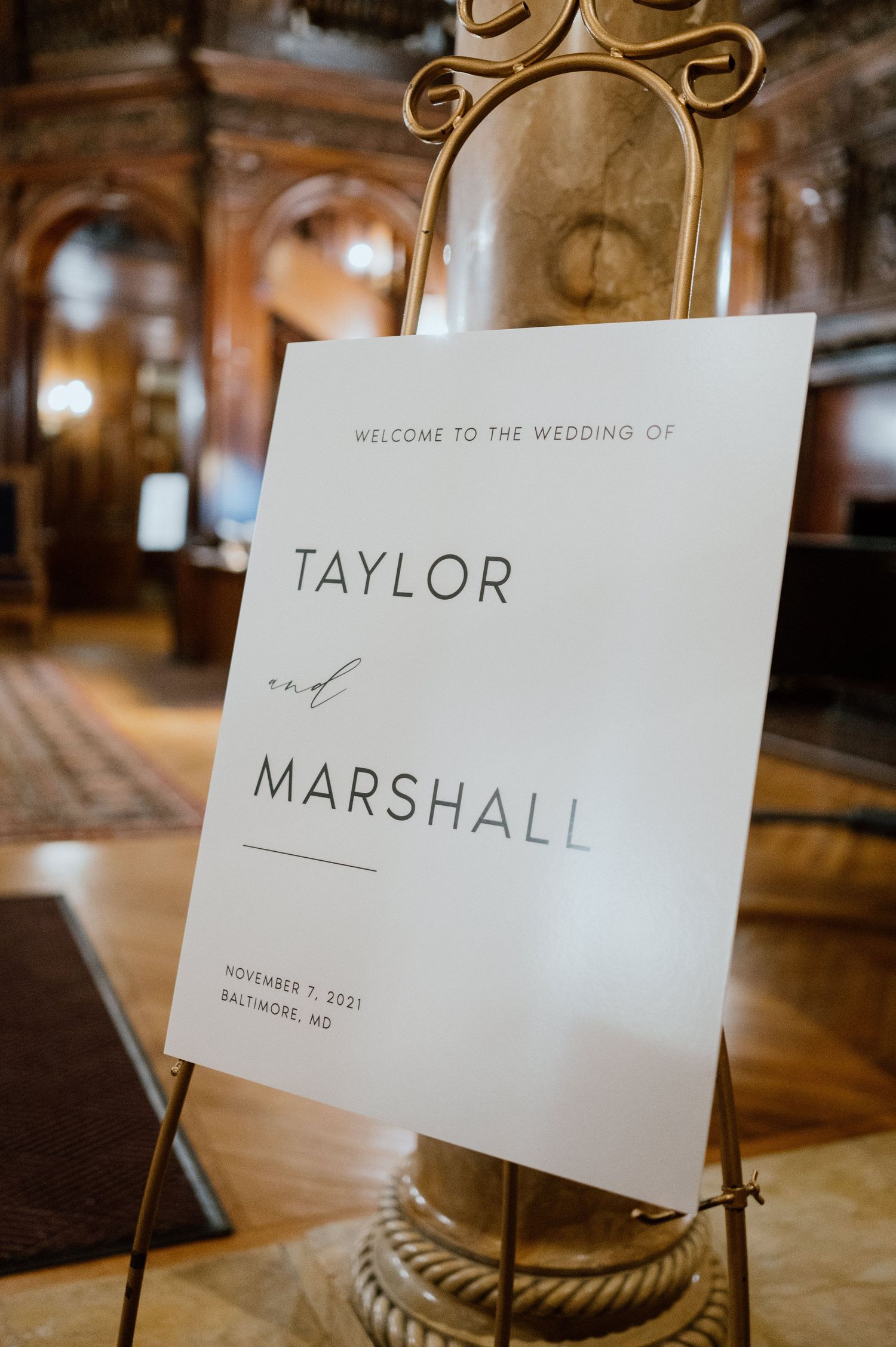 Wedding welcome sign for Taylor and Marshall displayed on gold stand in elegant venue.