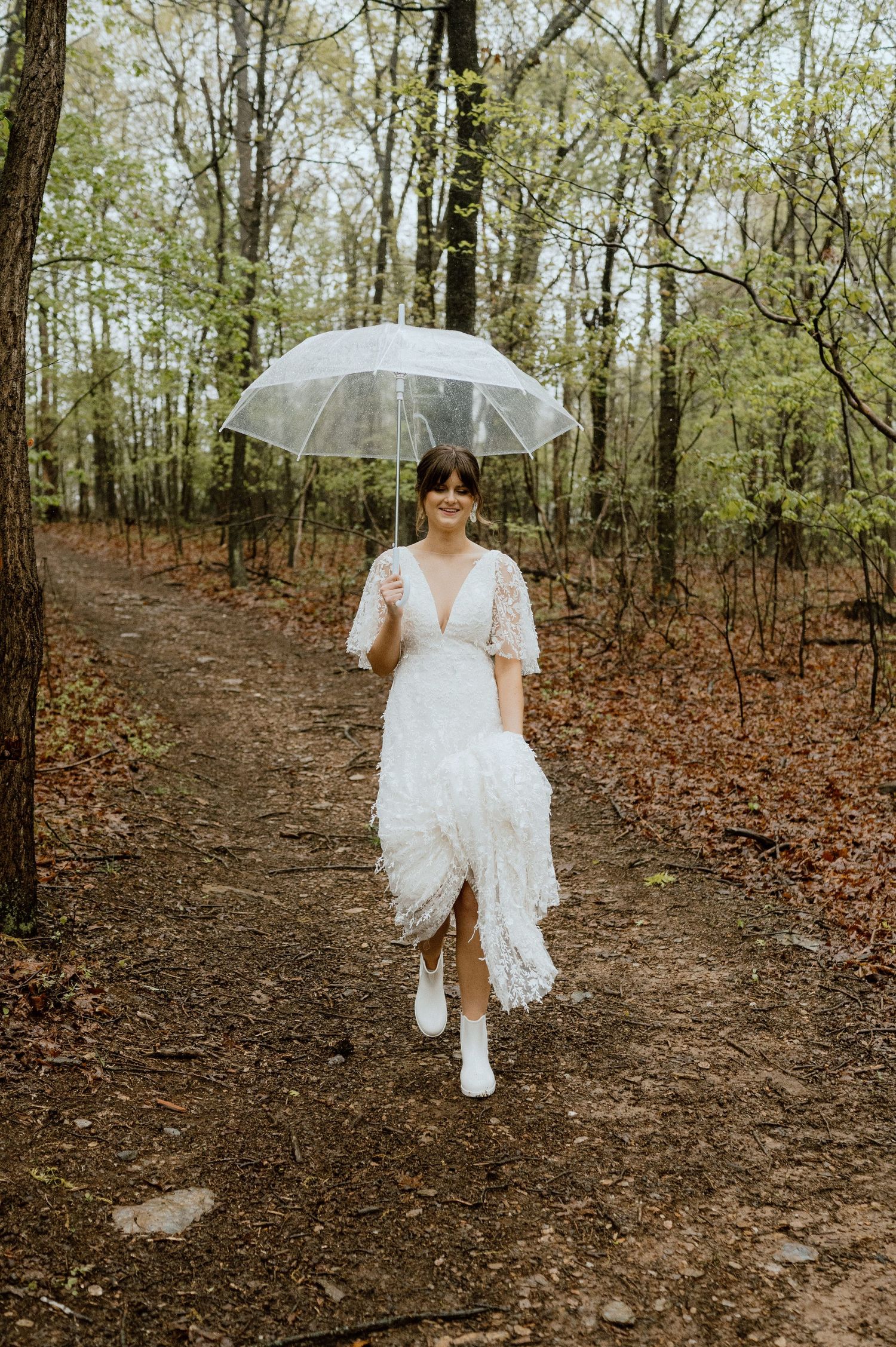 Bride in white dress walks forest path holding clear umbrella in autumn setting.