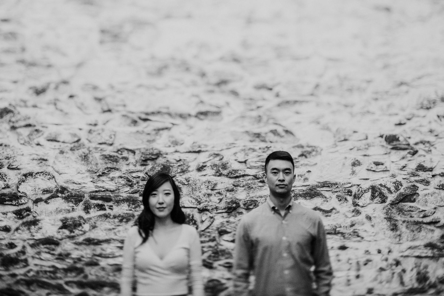 A black and white portrait of a couple standing against a textured stone wall.
