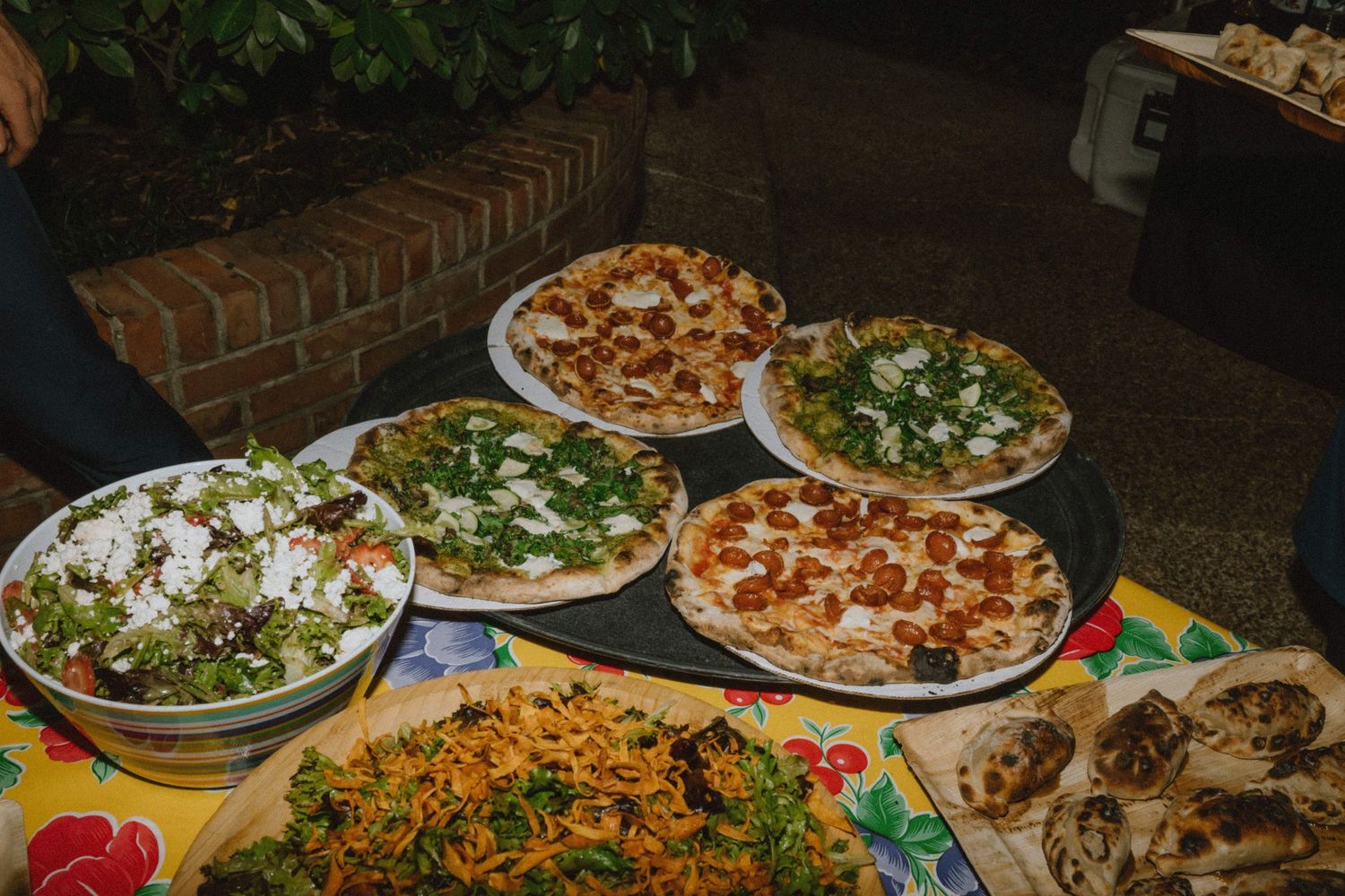 Various pizzas and salads displayed on a buffet table at an outdoor event.