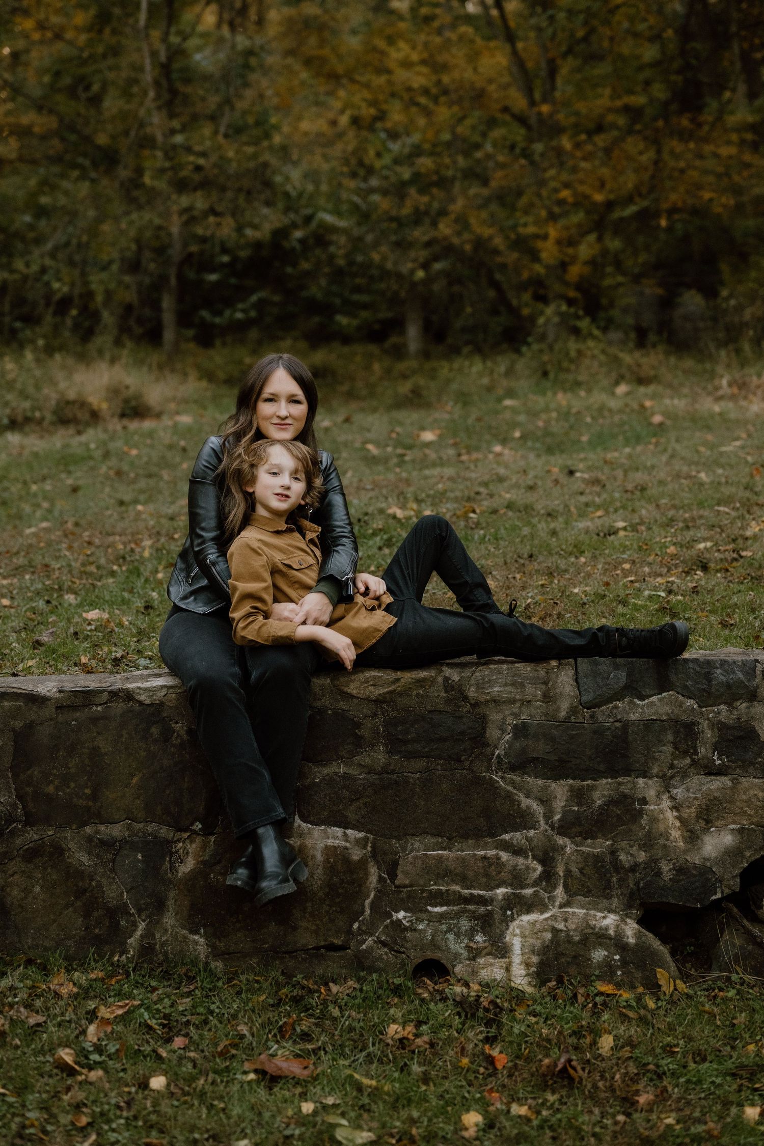 Two people sitting on a stone wall in an autumn setting wearing casual outfits.