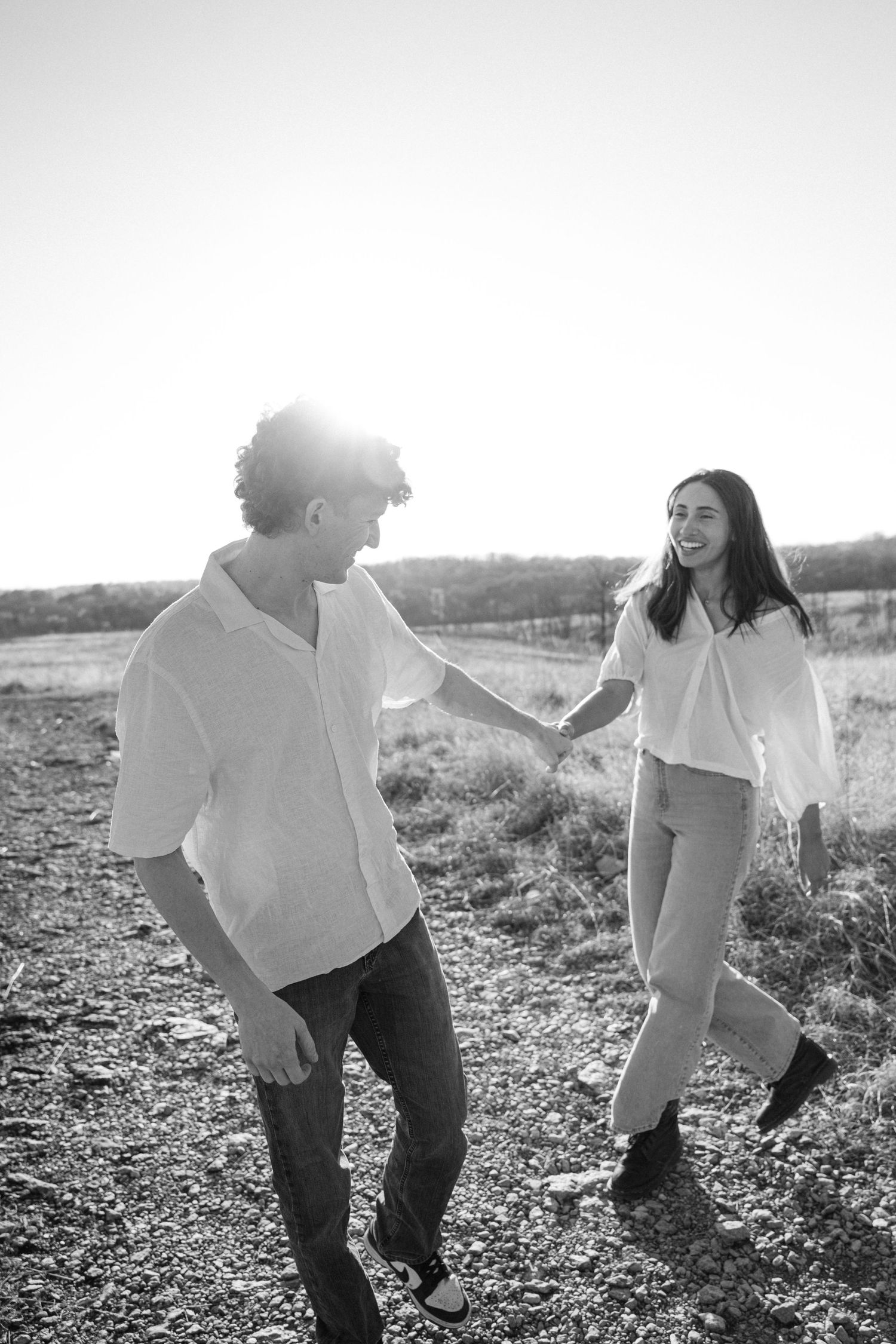 Black and white photo of a happy couple holding hands and walking on a rural path in golden sunlight.