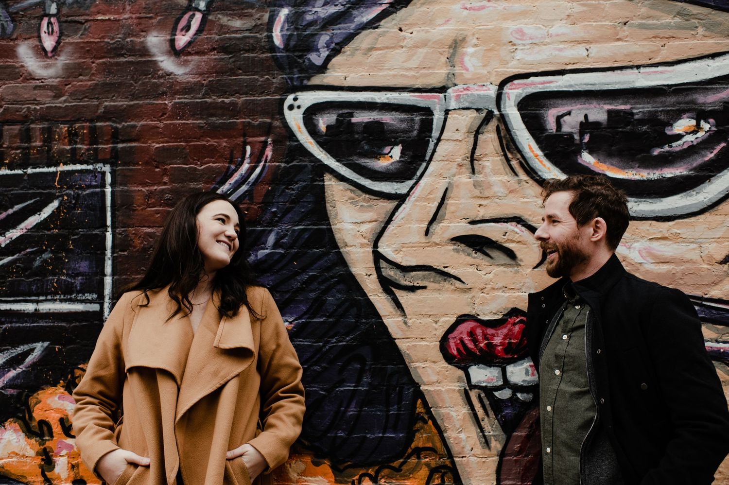 Couple smiling at each other in front of a colorful street art mural featuring a face with sunglasses.