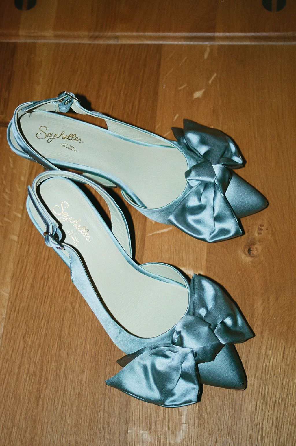 Light blue satin bridal shoes with bows displayed on wooden floor.