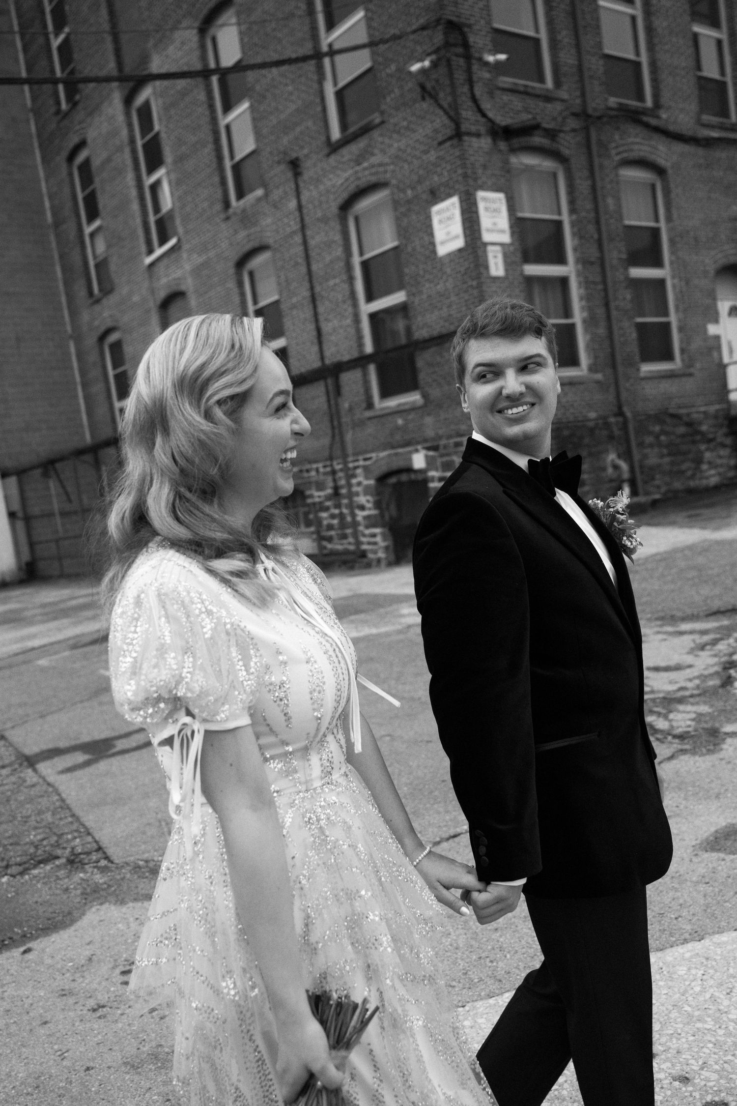 A couple in vintage-inspired wedding attire poses in front of a brick building.
