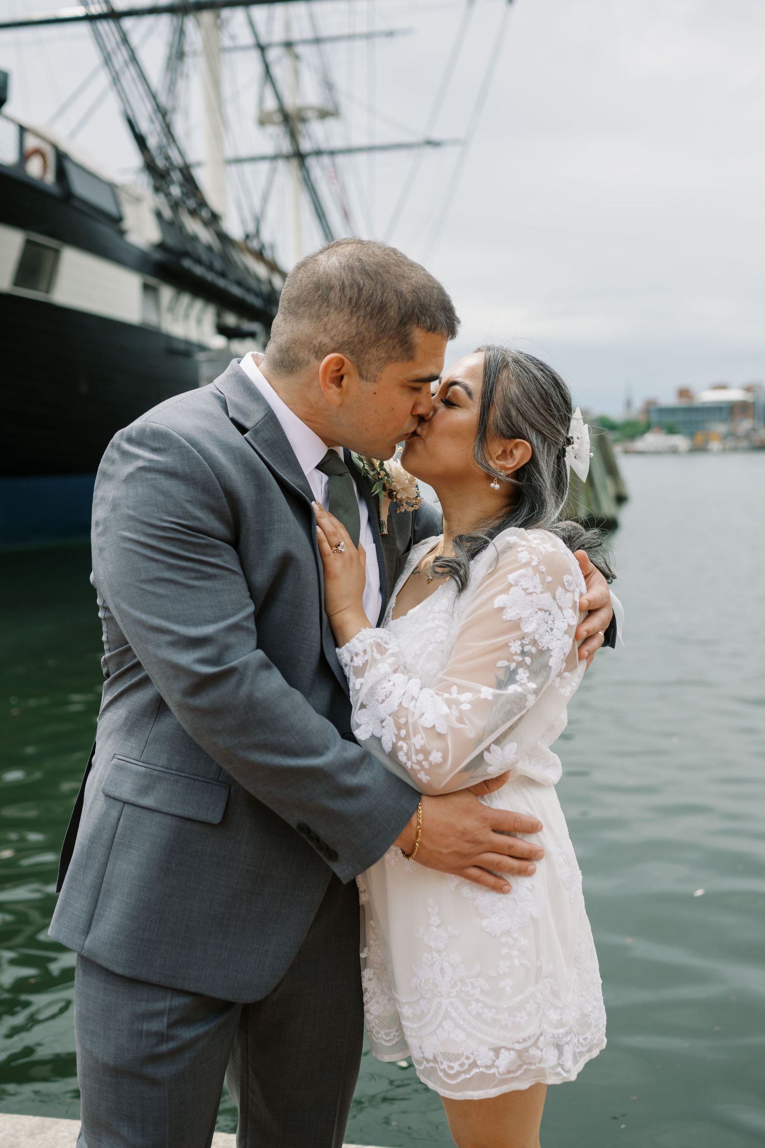 A romantic moment between newlyweds sharing a kiss by the water with a large ship docked in the background.