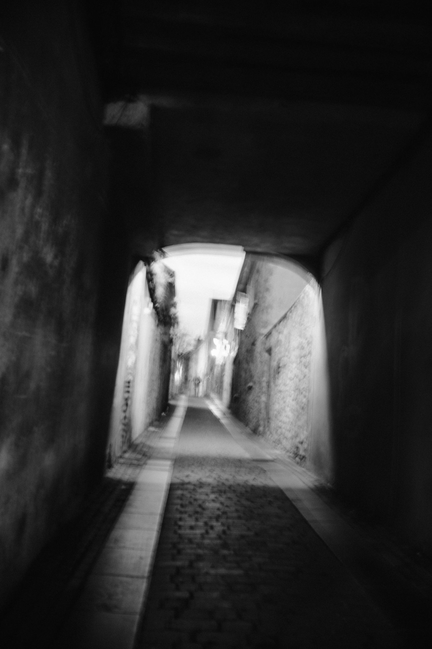 Moody black and white photograph of a narrow alleyway through an arched tunnel.