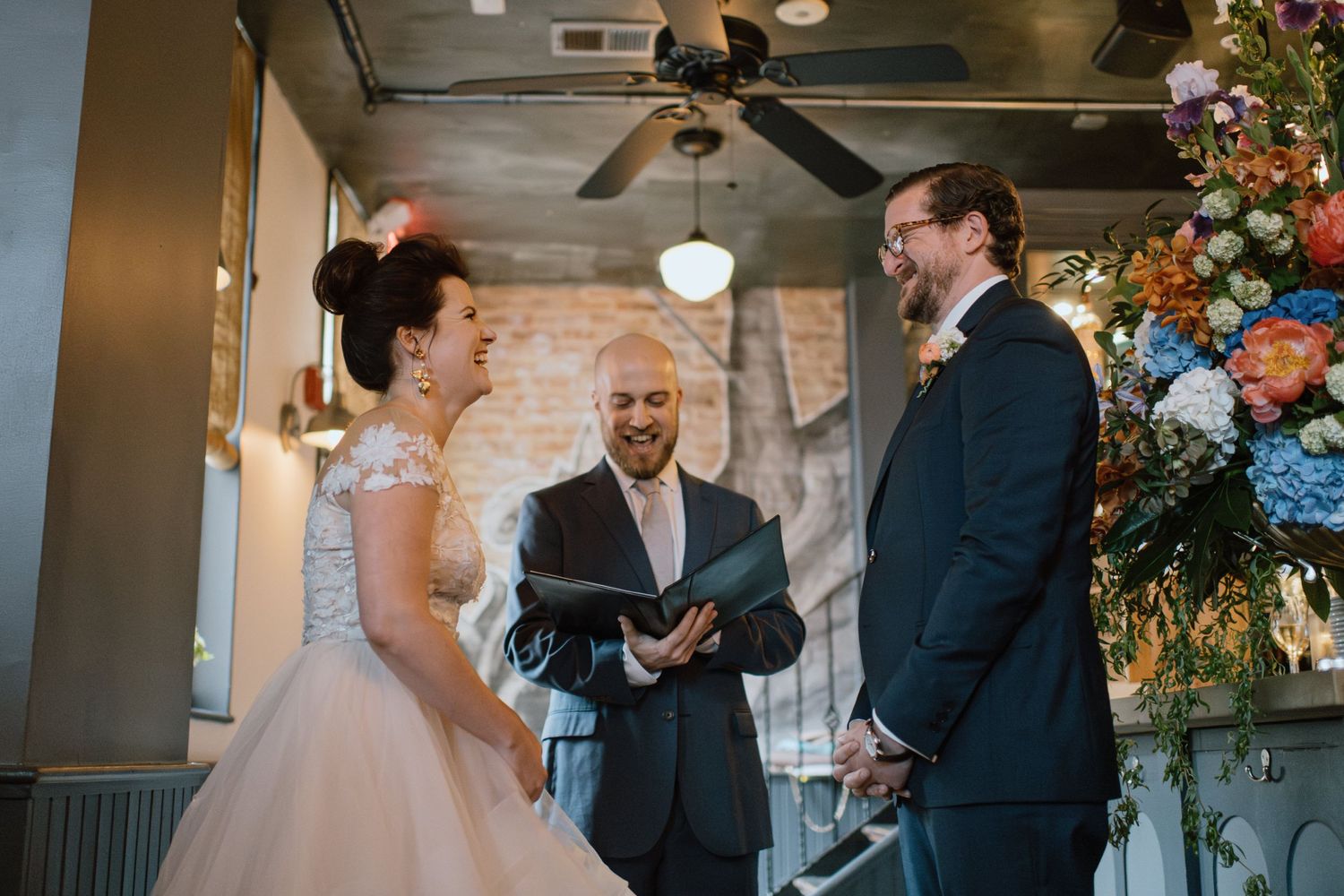 A wedding ceremony takes place in an intimate indoor venue with a colorful floral arrangement to the side.