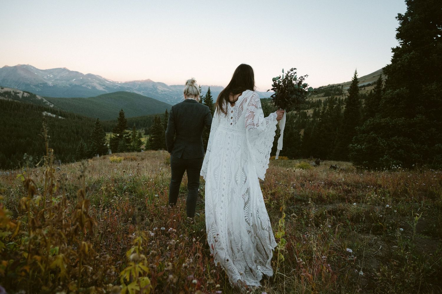 A couple walking through mountain wildflowers at sunset while wearing formal attire.