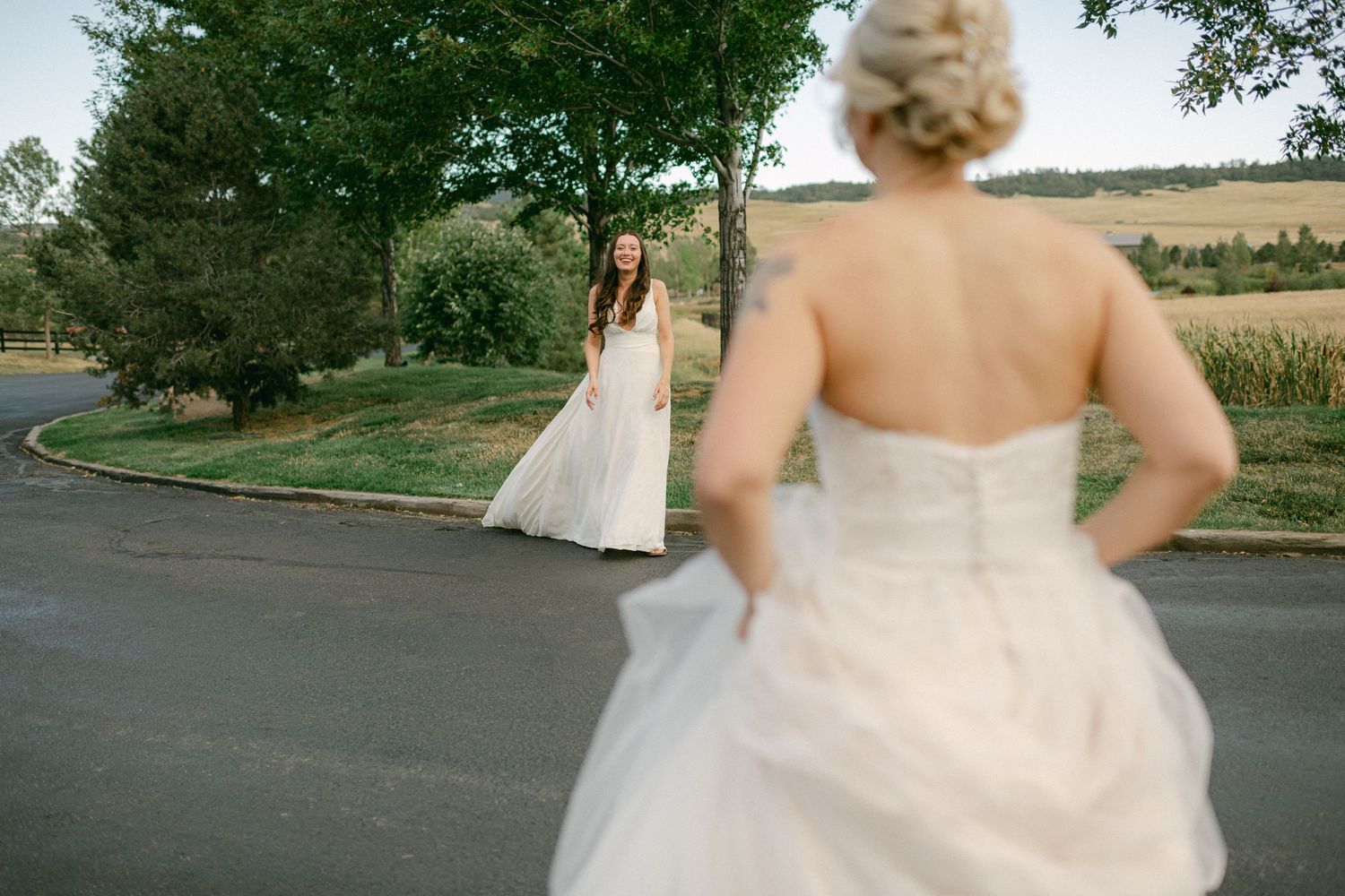 Two people in wedding dresses stand on a winding road surrounded by countryside.