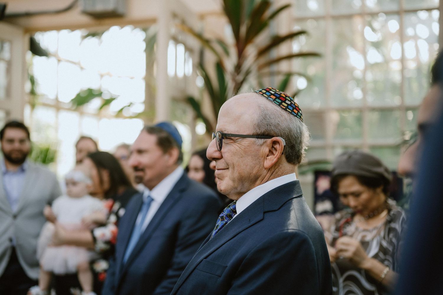 Wedding guests gather in a sunlit conservatory with tropical plants.