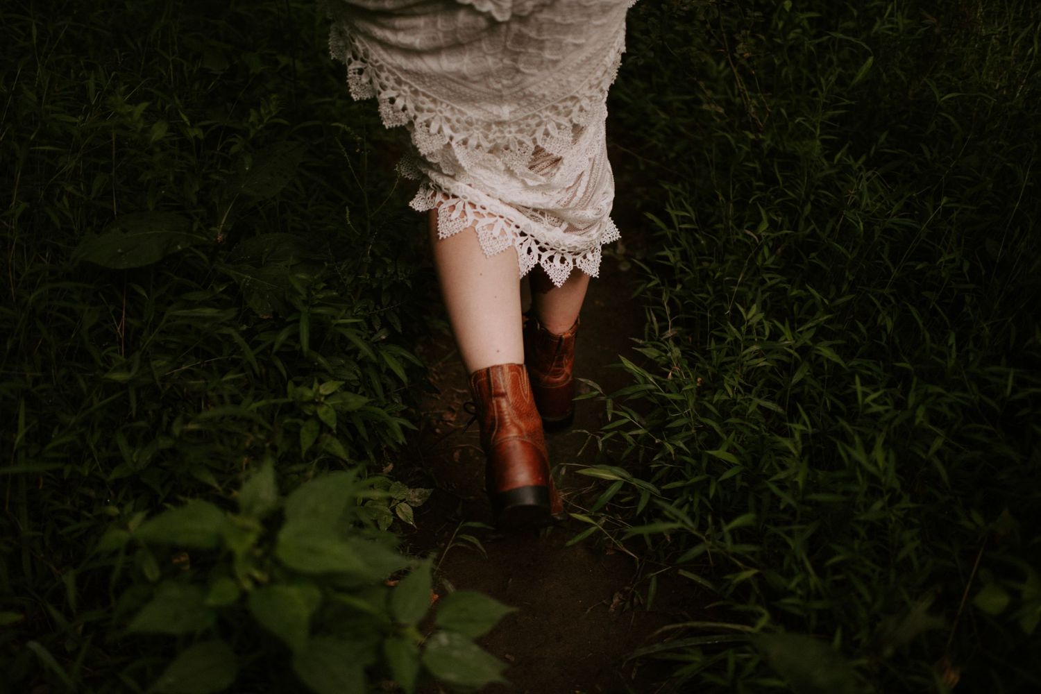 Brown leather boots contrast with delicate white lace dress hem in dark grass.
